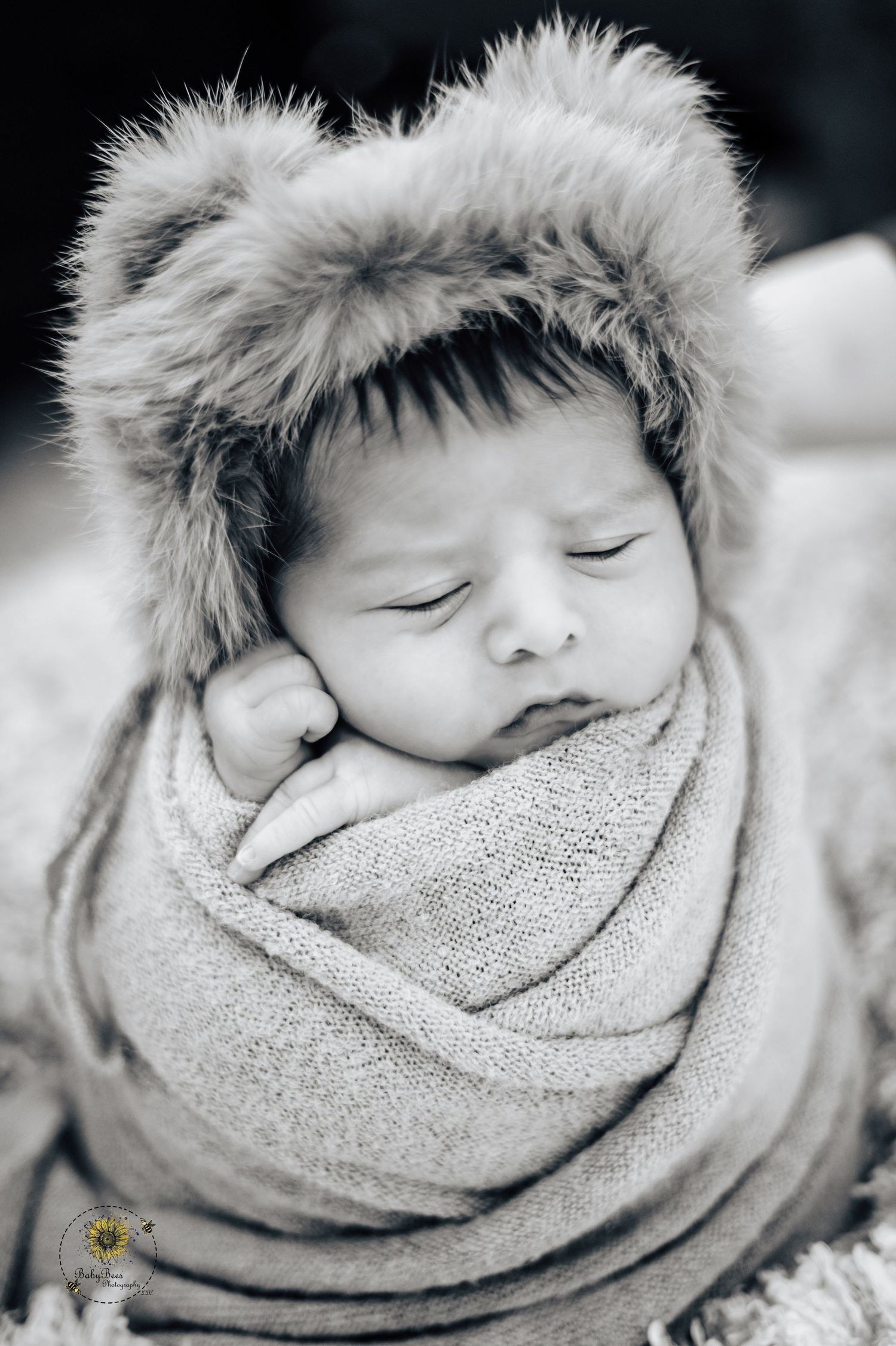 A sleeping newborn wrapped in a soft gray blanket with a fur-trimmed hat in black and white photography.