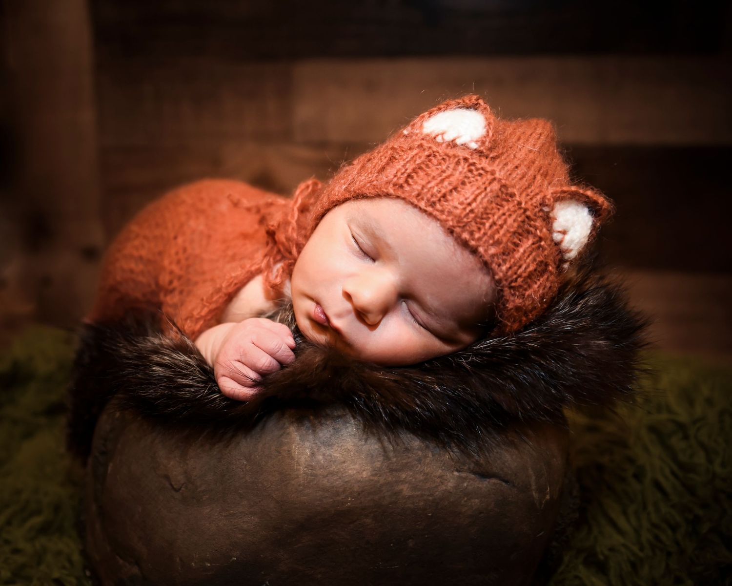 Newborn baby wearing orange knit hat sleeps peacefully on soft green moss textured backdrop.