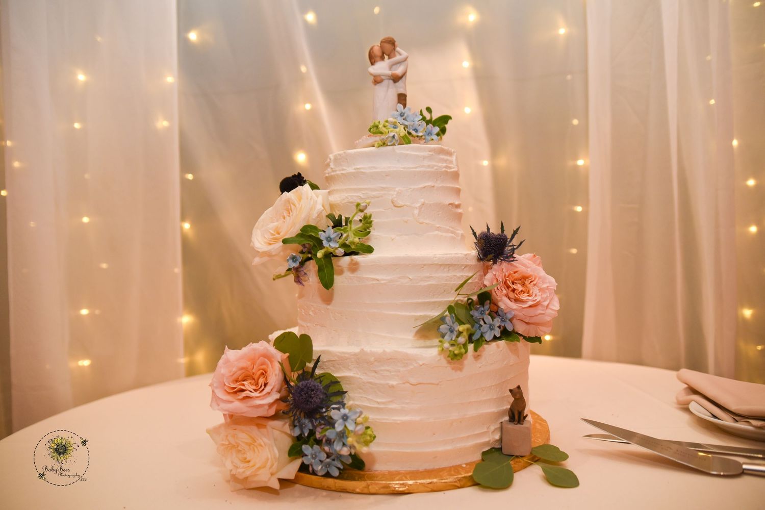 Elegant white tiered wedding cake decorated with peach roses and blue flowers against a backdrop of twinkling lights.