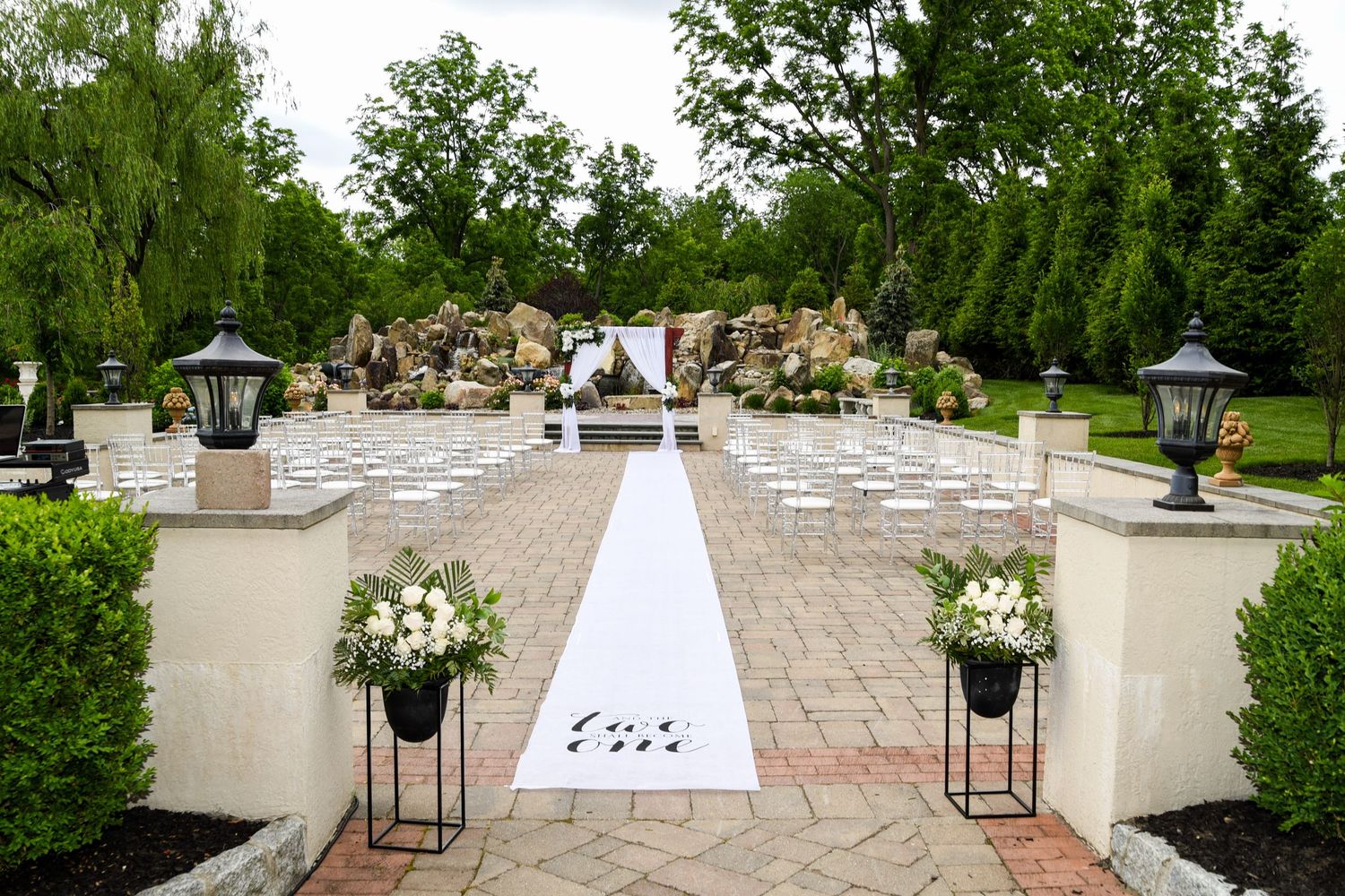 Outdoor wedding ceremony setup with white aisle runner and floral arrangements leading to stone garden backdrop.