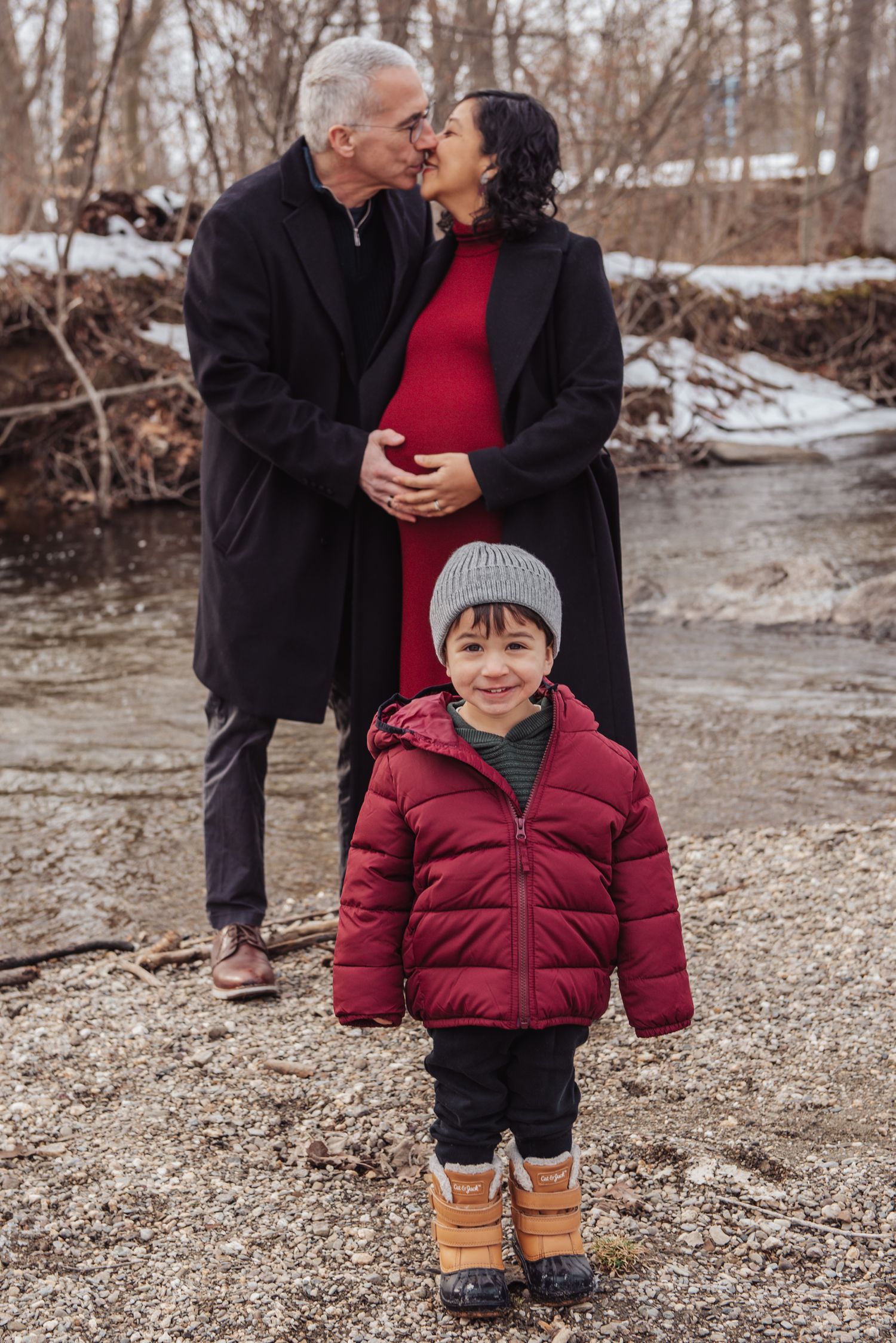 A young child in a red winter coat smiles while walking outdoors with two adults on a snowy winter day.
