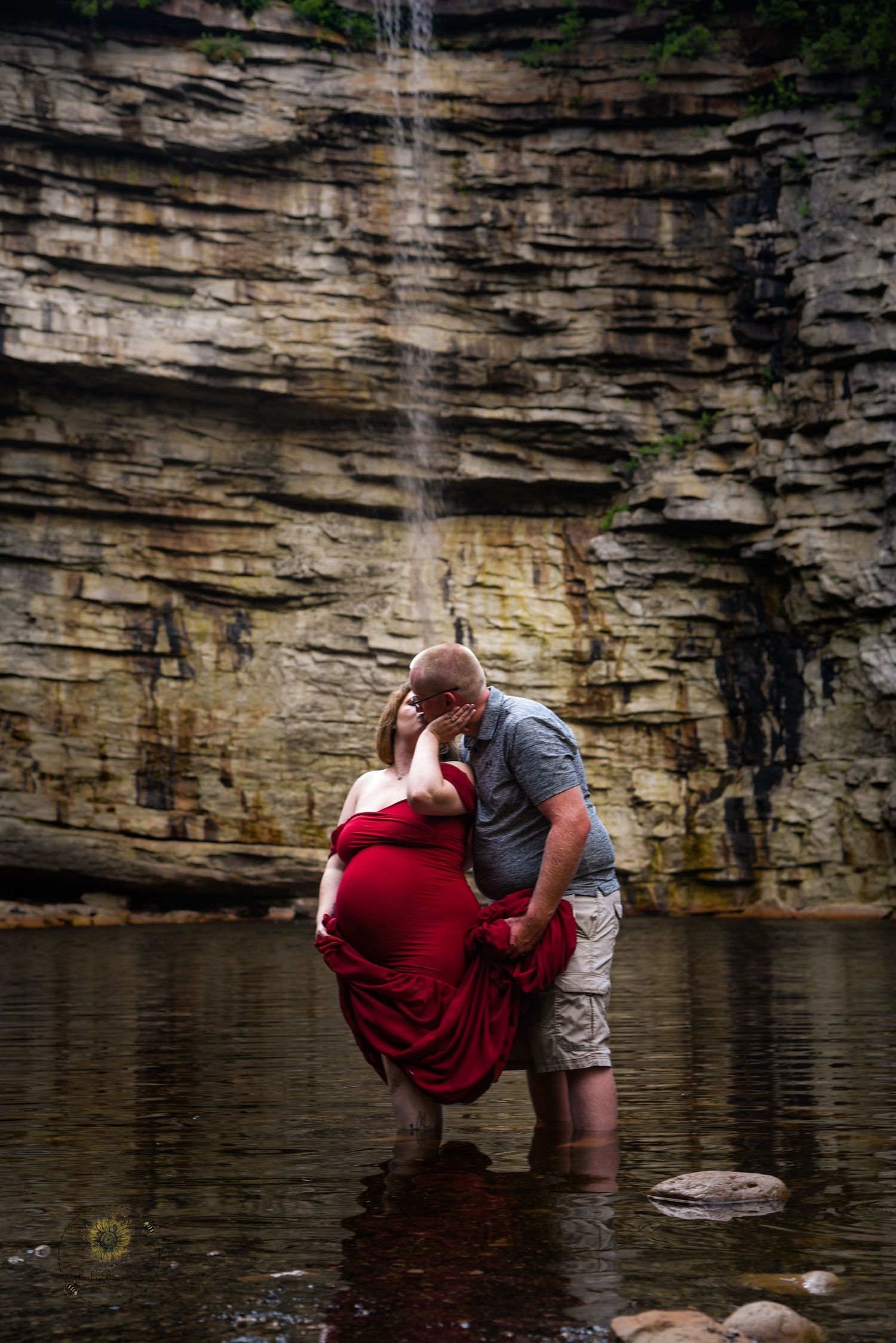 A couple embraces in front of a dramatic limestone cliff wall beside a tranquil lake.