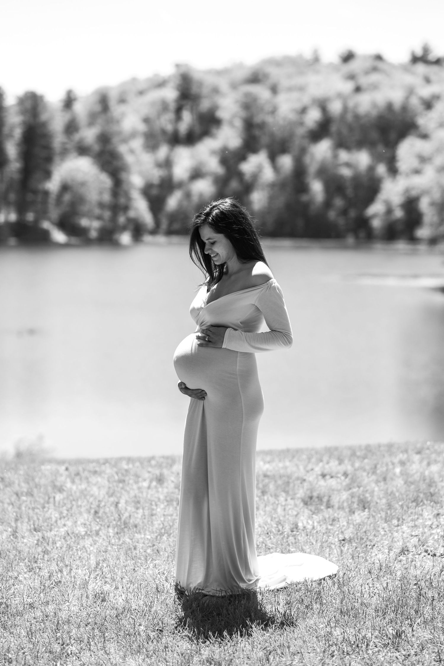 A black and white maternity photo shows an expectant mother in a flowing dress standing by a serene lake.