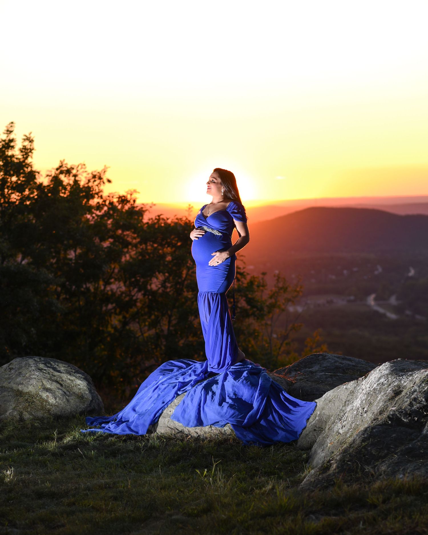 A figure in a flowing blue dress poses dramatically on rocky terrain against a vibrant sunset backdrop.