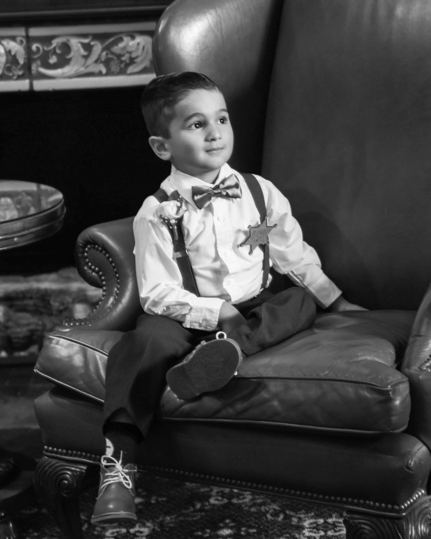 Black and white portrait of young ring bearer lounging on leather chair at formal event.