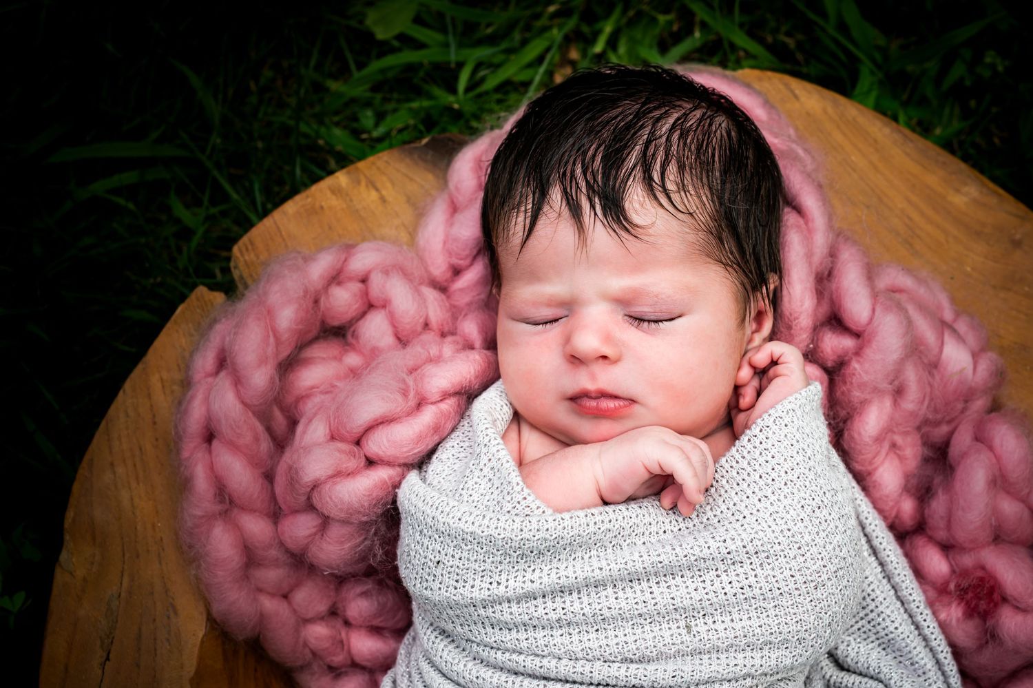 Sleeping newborn wrapped in white swaddle rests peacefully on pink chunky knit blanket in wooden bowl.