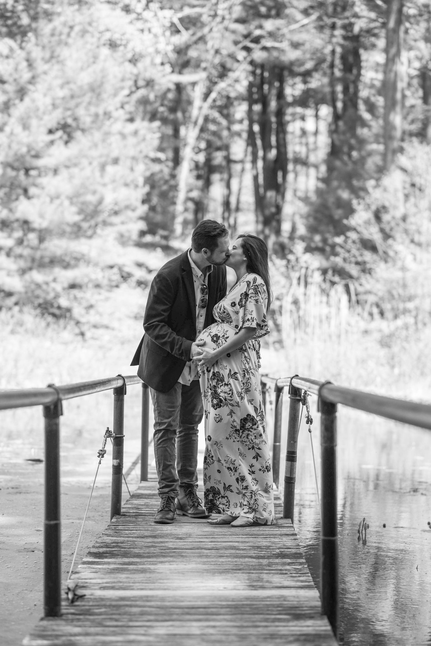 A romantic couple shares tender moments on a wooden dock surrounded by trees in black and white photography.