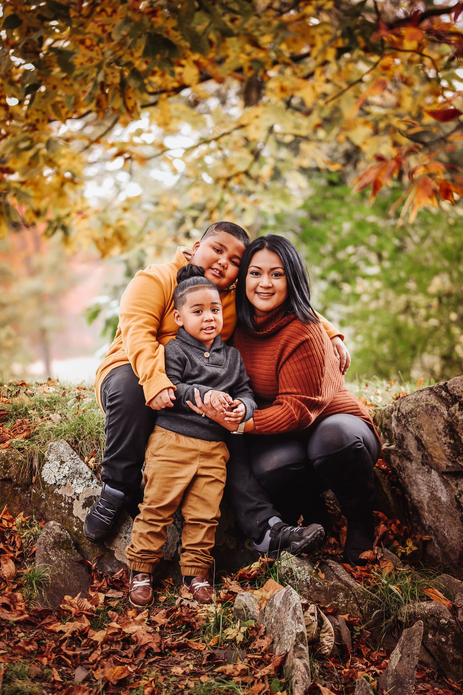 A family sits together on rocks surrounded by autumn foliage with golden leaves overhead in a park setting.