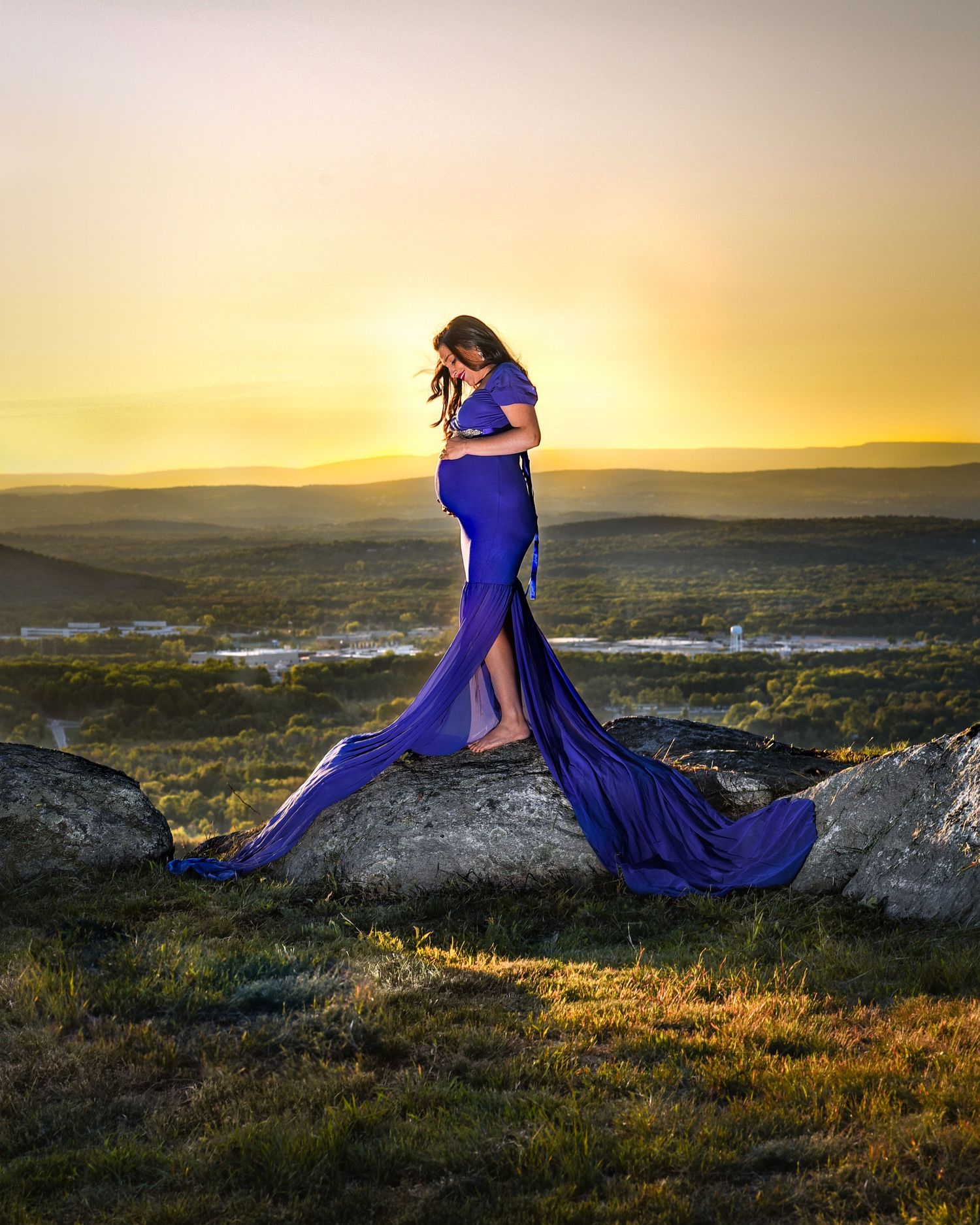 A figure in a flowing royal blue gown sits atop a rocky overlook at sunset with mountains and valleys in the distance.