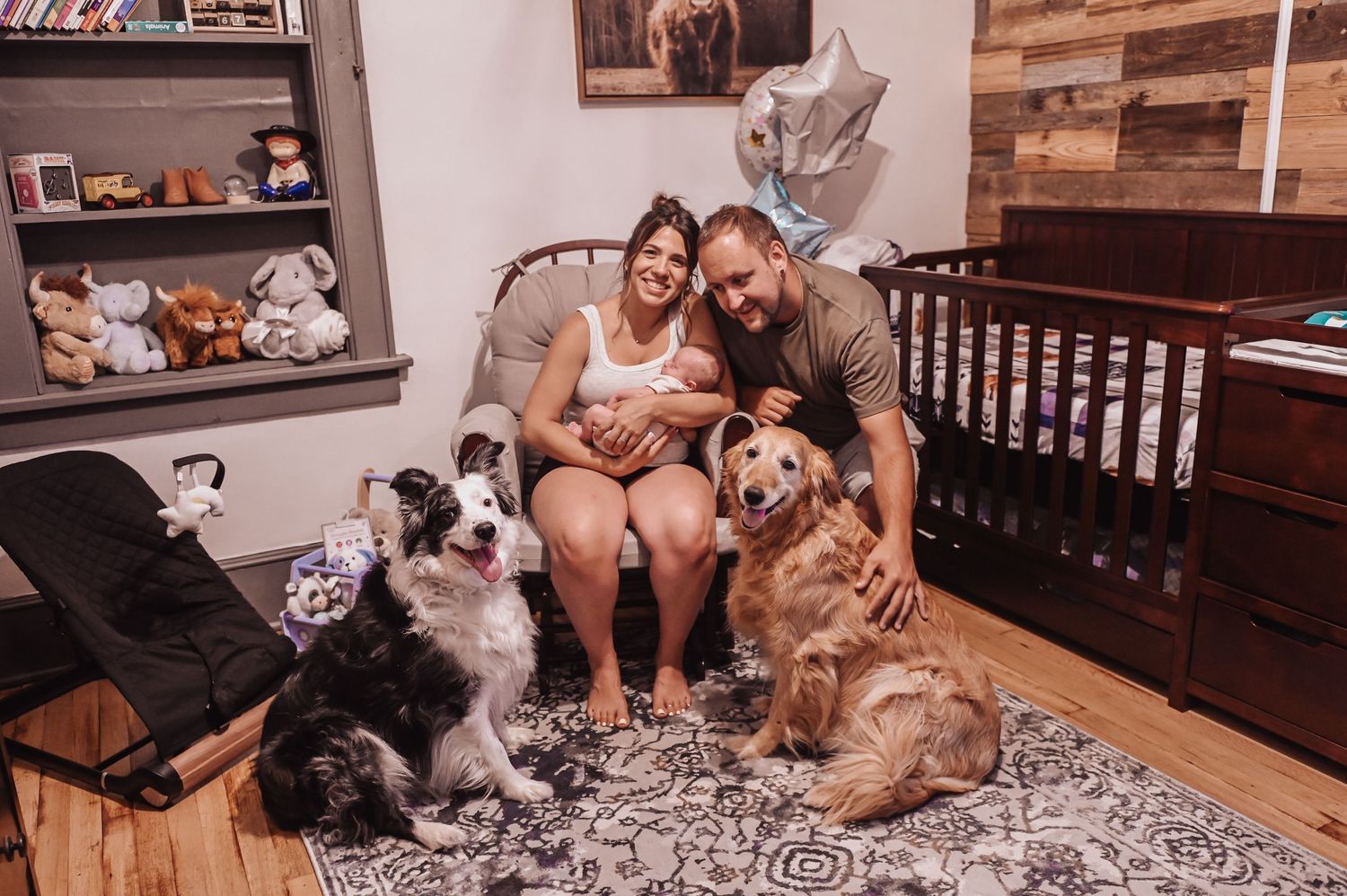 A couple sits on a patterned rug in a nursery room with three dogs, surrounded by baby furniture and decor.