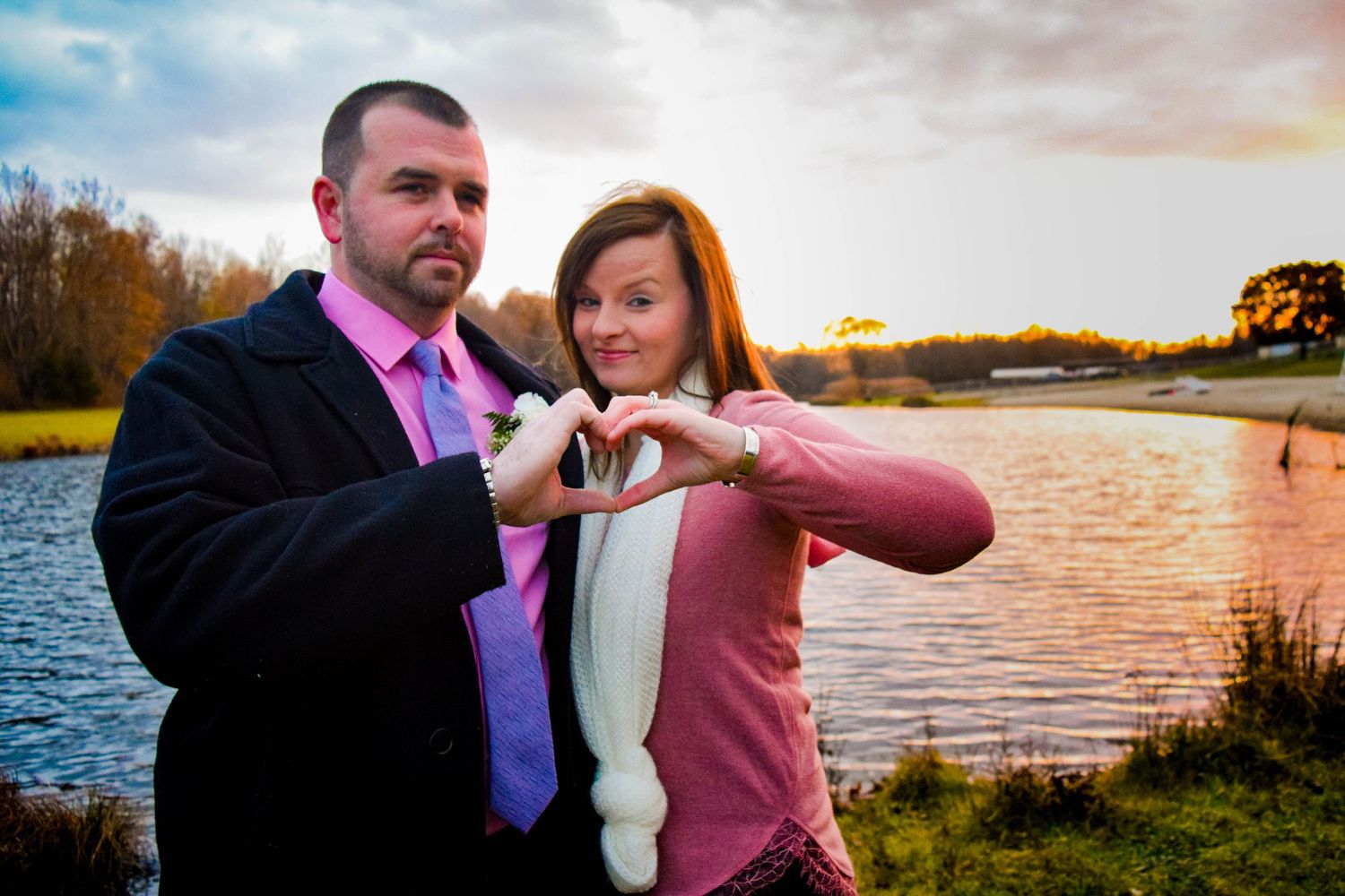 A couple forms a heart shape with their hands against a sunset lake backdrop.