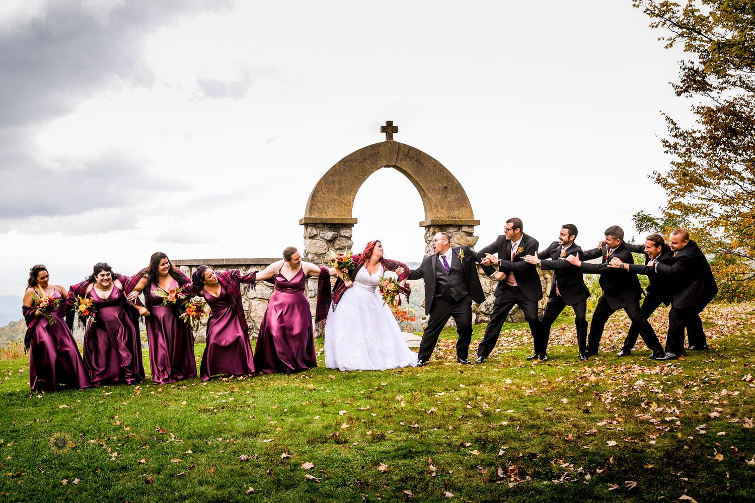 Wedding group photo with bridal party arranged on grassy hill near stone arch.