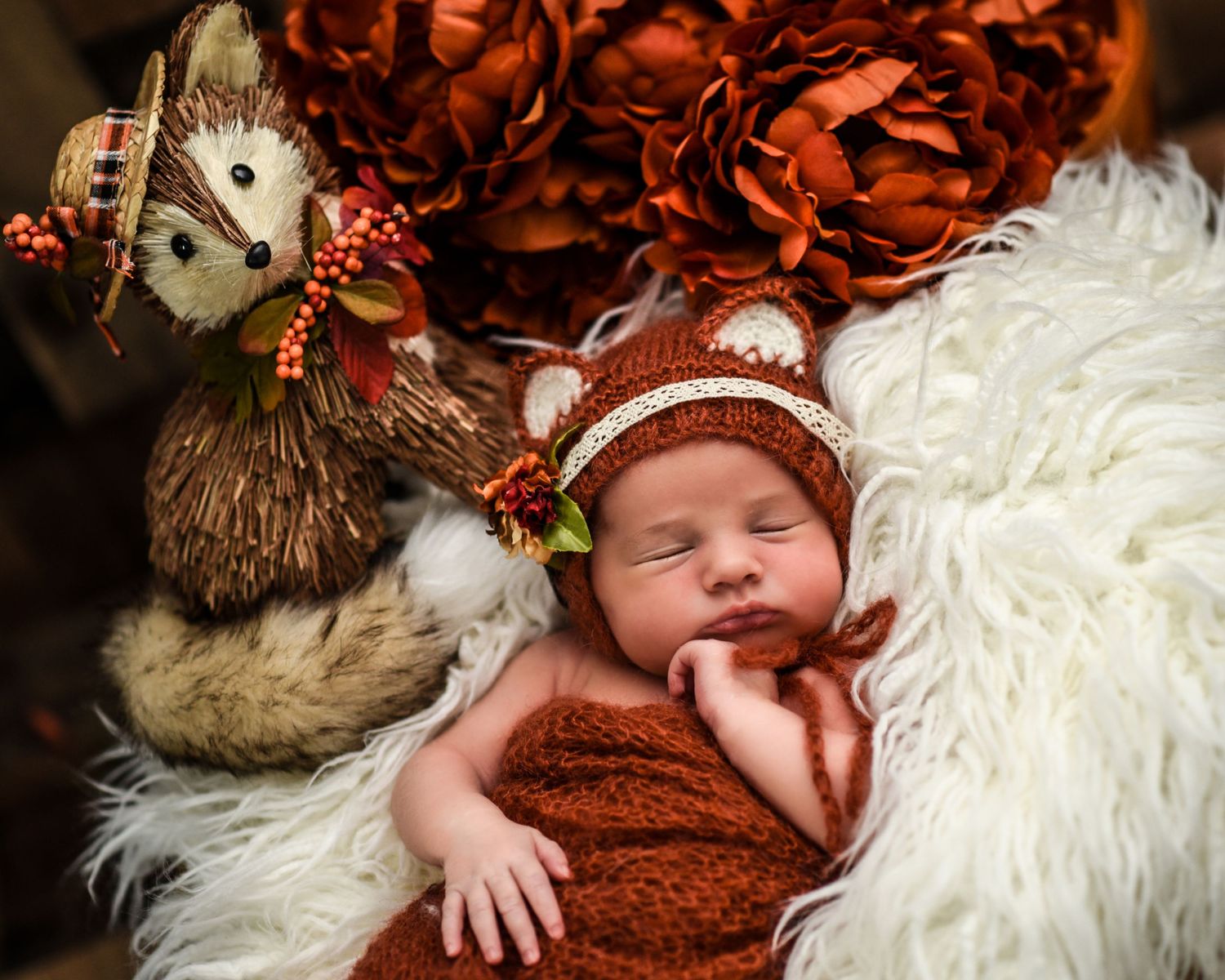 A newborn wearing a brown knit outfit and floral headpiece lies on soft white fur during a professional photo session.