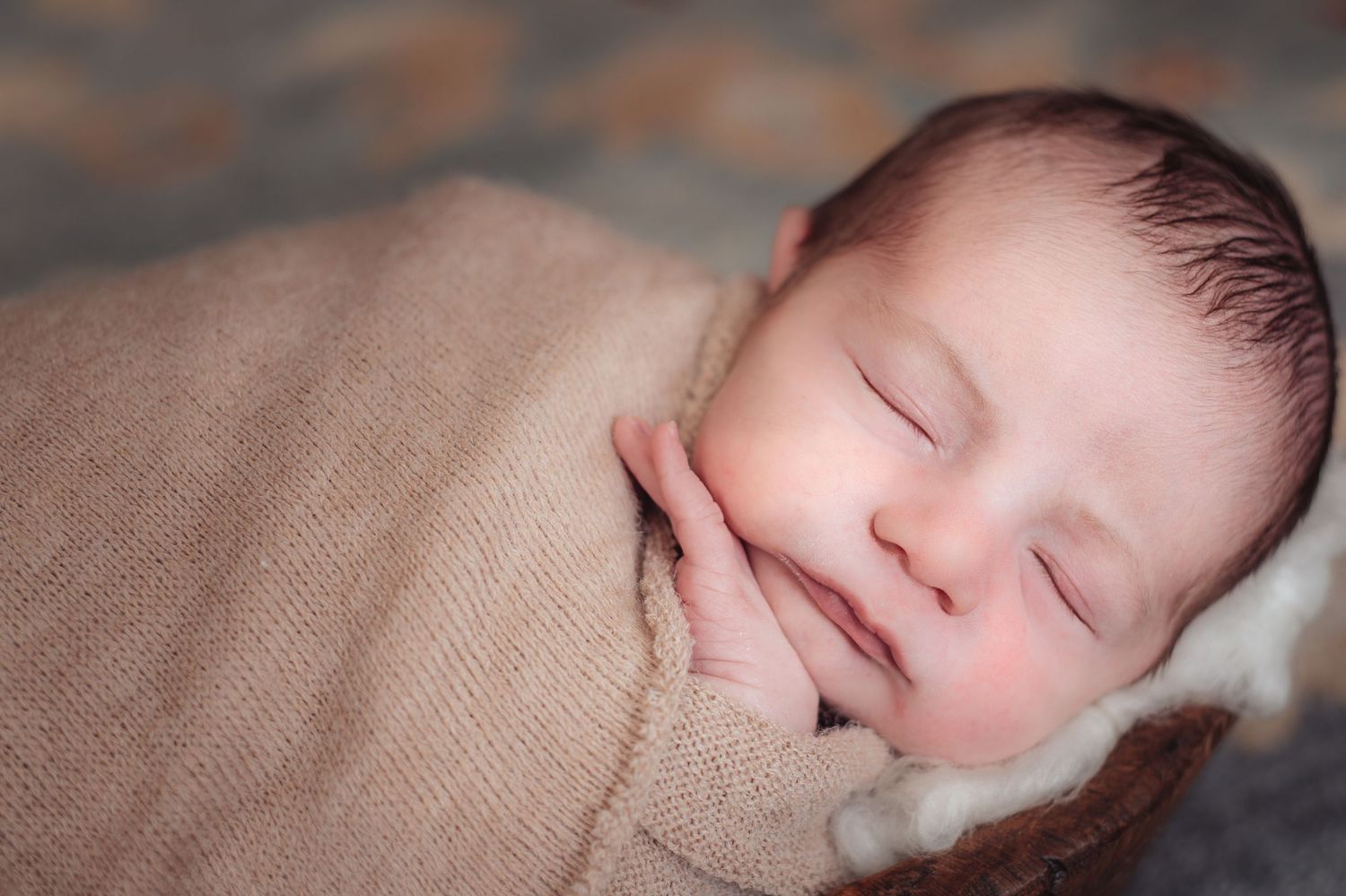 Sleeping newborn baby wrapped in soft beige blanket with peaceful expression during professional photoshoot.