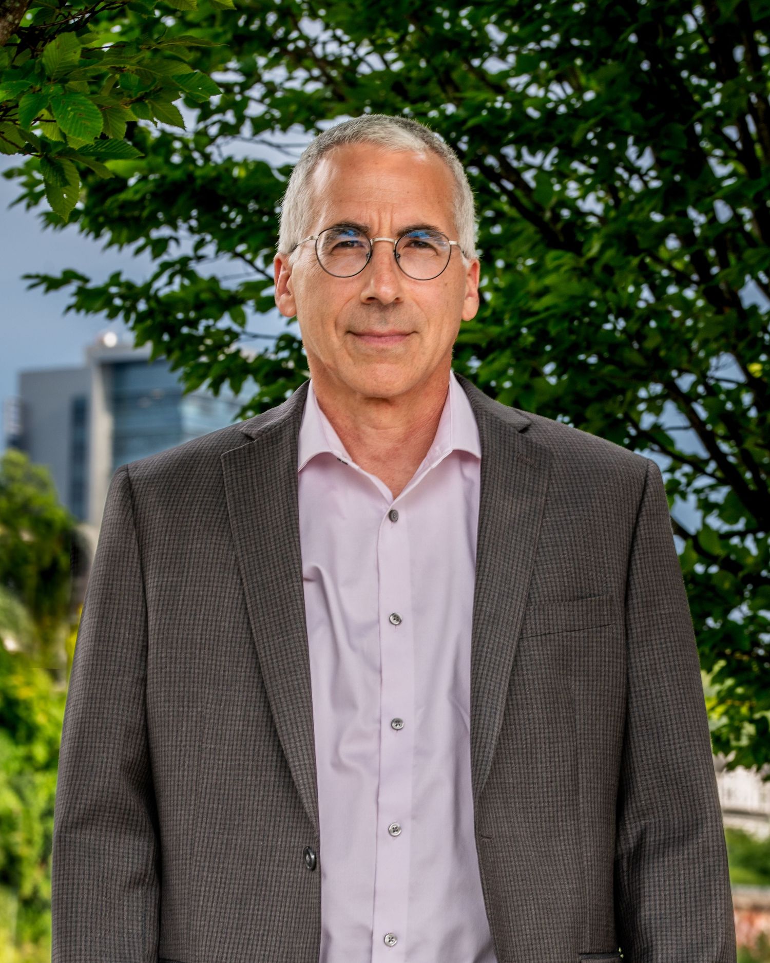 Professional headshot portrait of someone wearing glasses, a pink dress shirt and gray suit jacket in an outdoor setting.