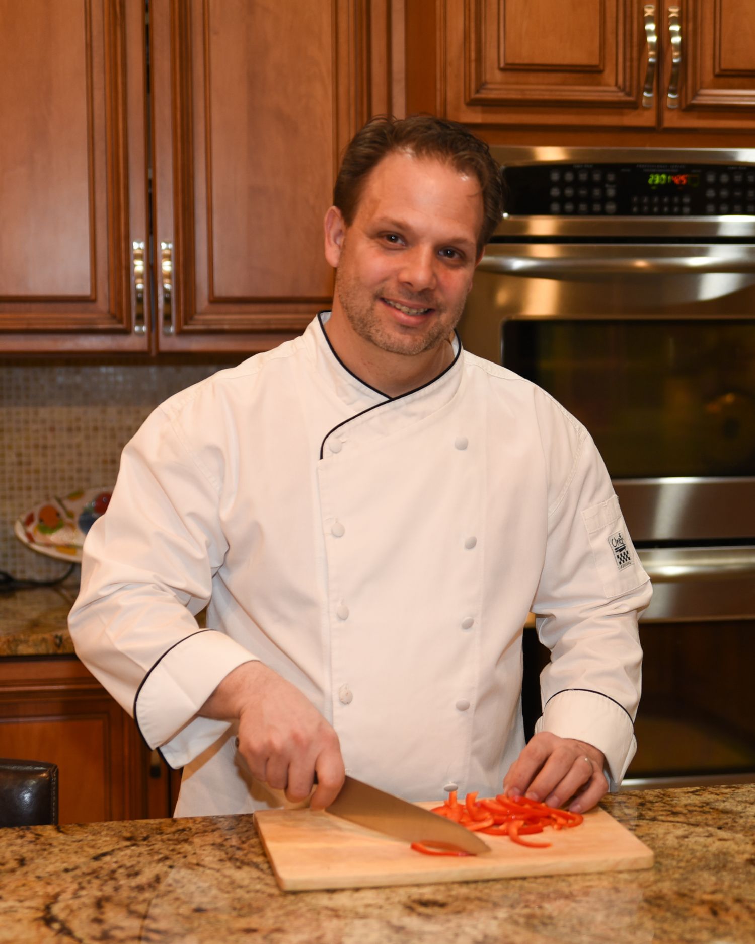 Chef in white uniform chopping red peppers on wooden cutting board in upscale kitchen with granite counters.