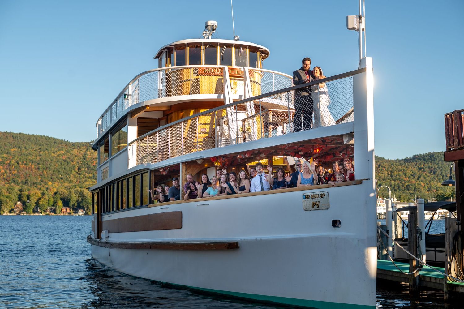A classic wooden tour boat filled with wedding guests cruises across a scenic lake.