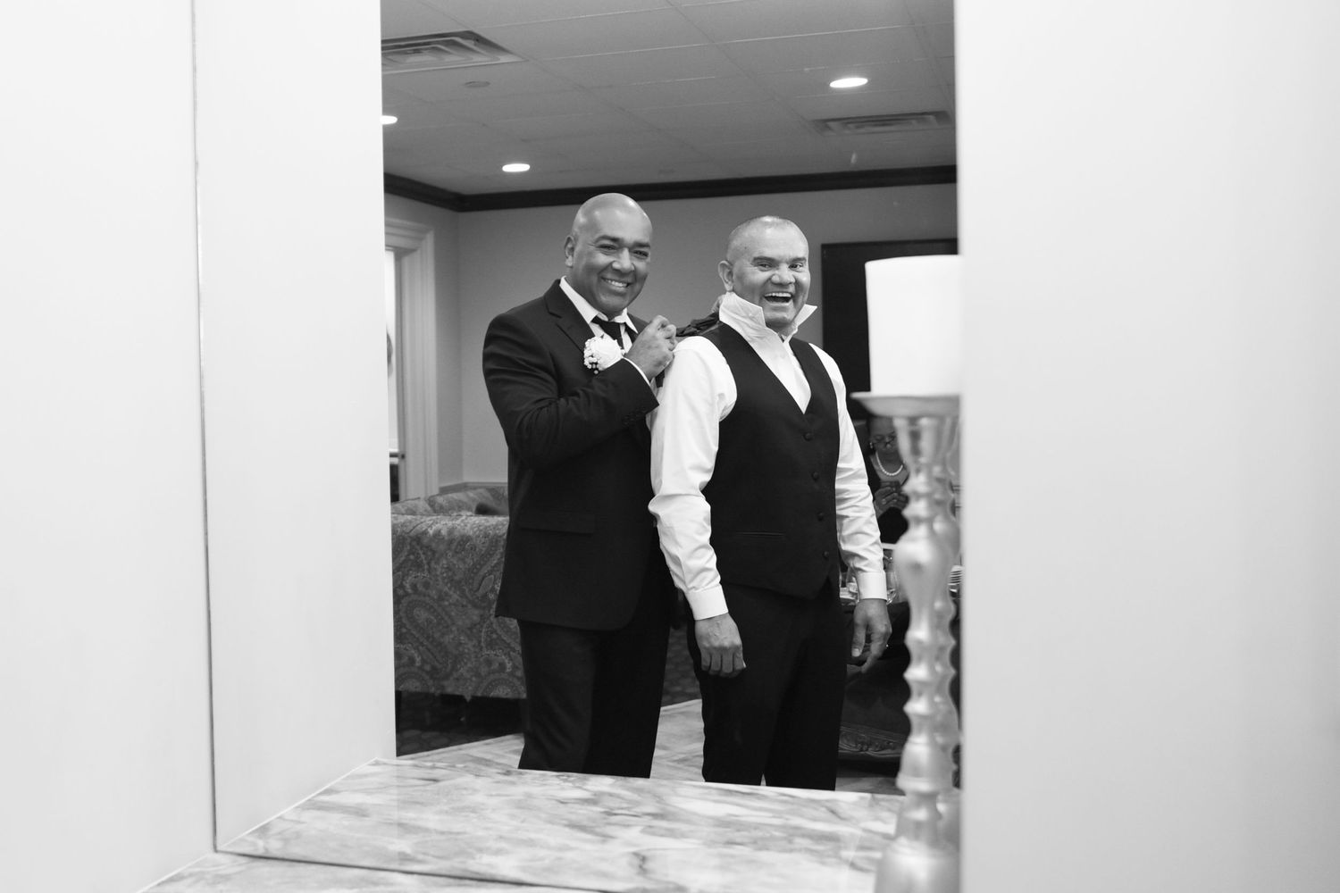 Black and white photo of two men in formal attire smiling in mirror reflection.