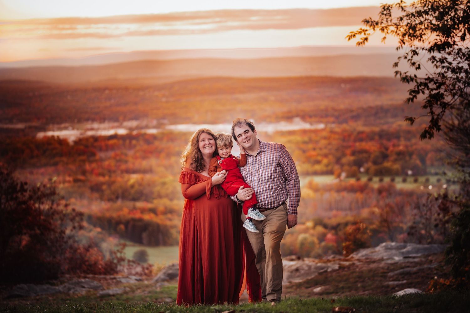 Couple in red dress and plaid shirt embrace during golden sunset over rolling hills in autumn landscape.