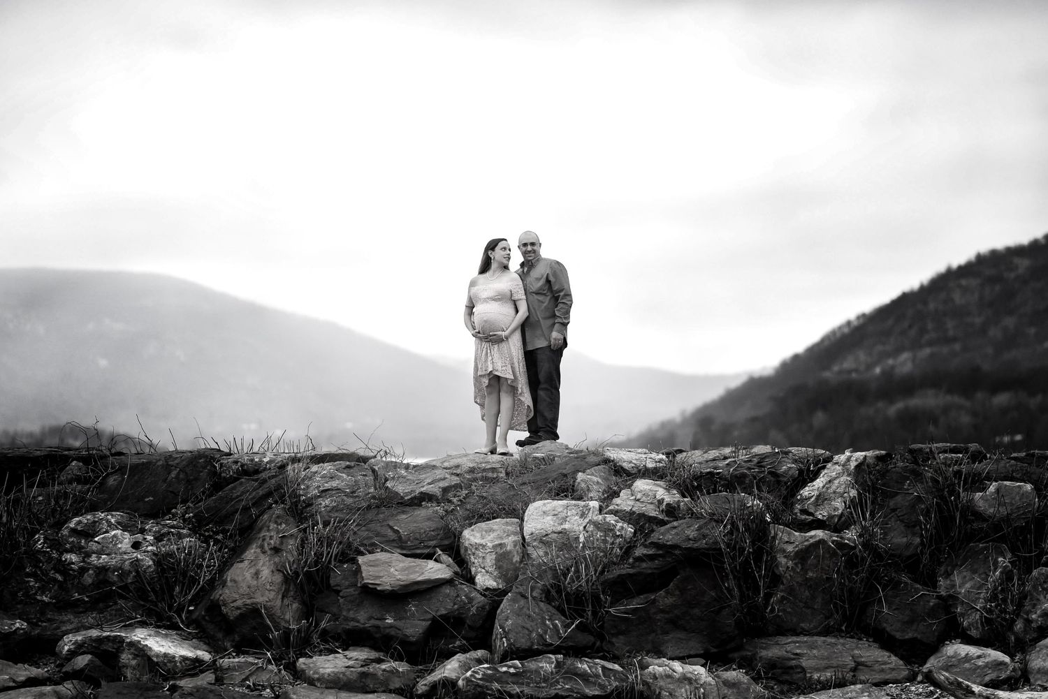 A couple stands together on a rocky outcrop overlooking misty mountain valleys in black and white.