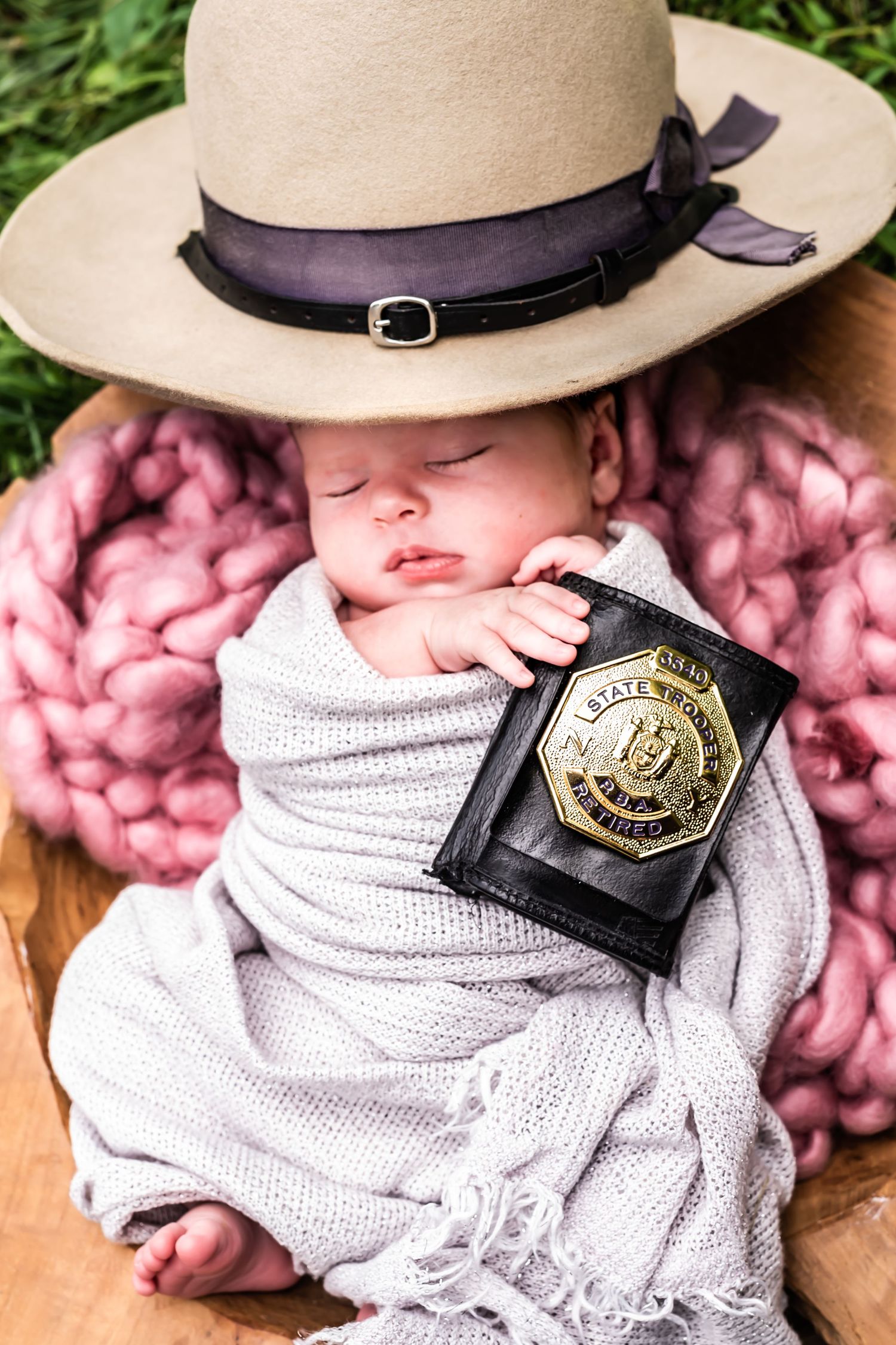 A baby wearing a cowboy hat and white swaddle blanket lies next to a police badge on a pink textured background.