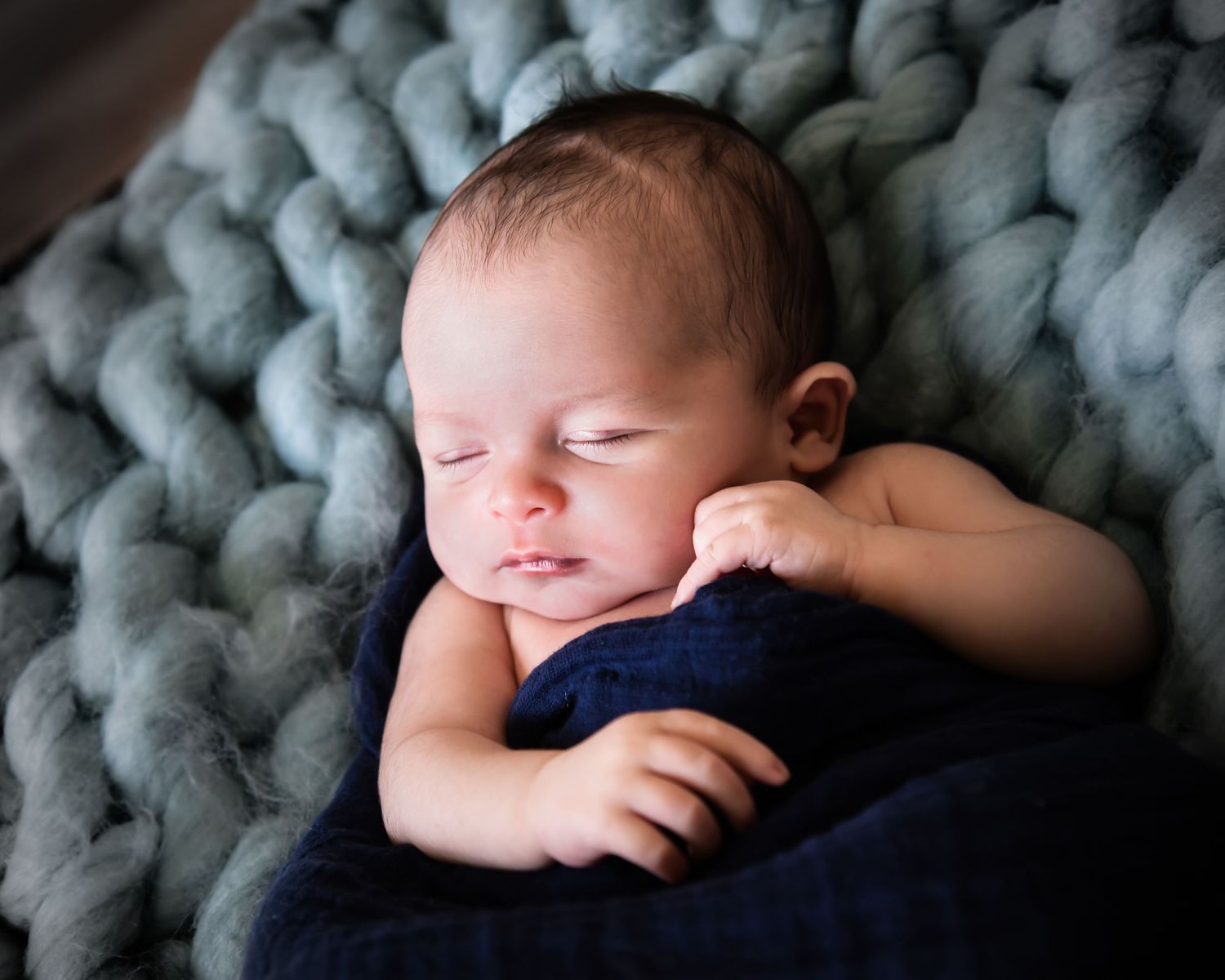 Sleeping newborn baby rests peacefully on chunky knit gray blanket in soft natural light.