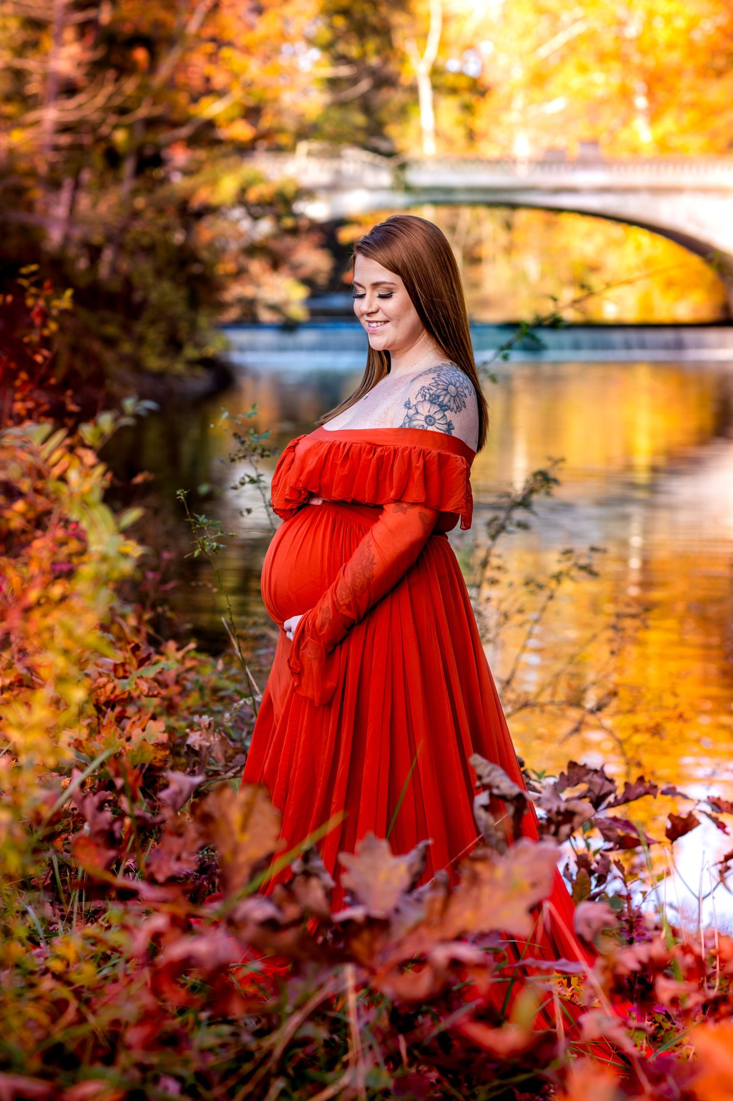 A person in a flowing red dress poses by a lake during autumn with golden foliage reflecting in the water.