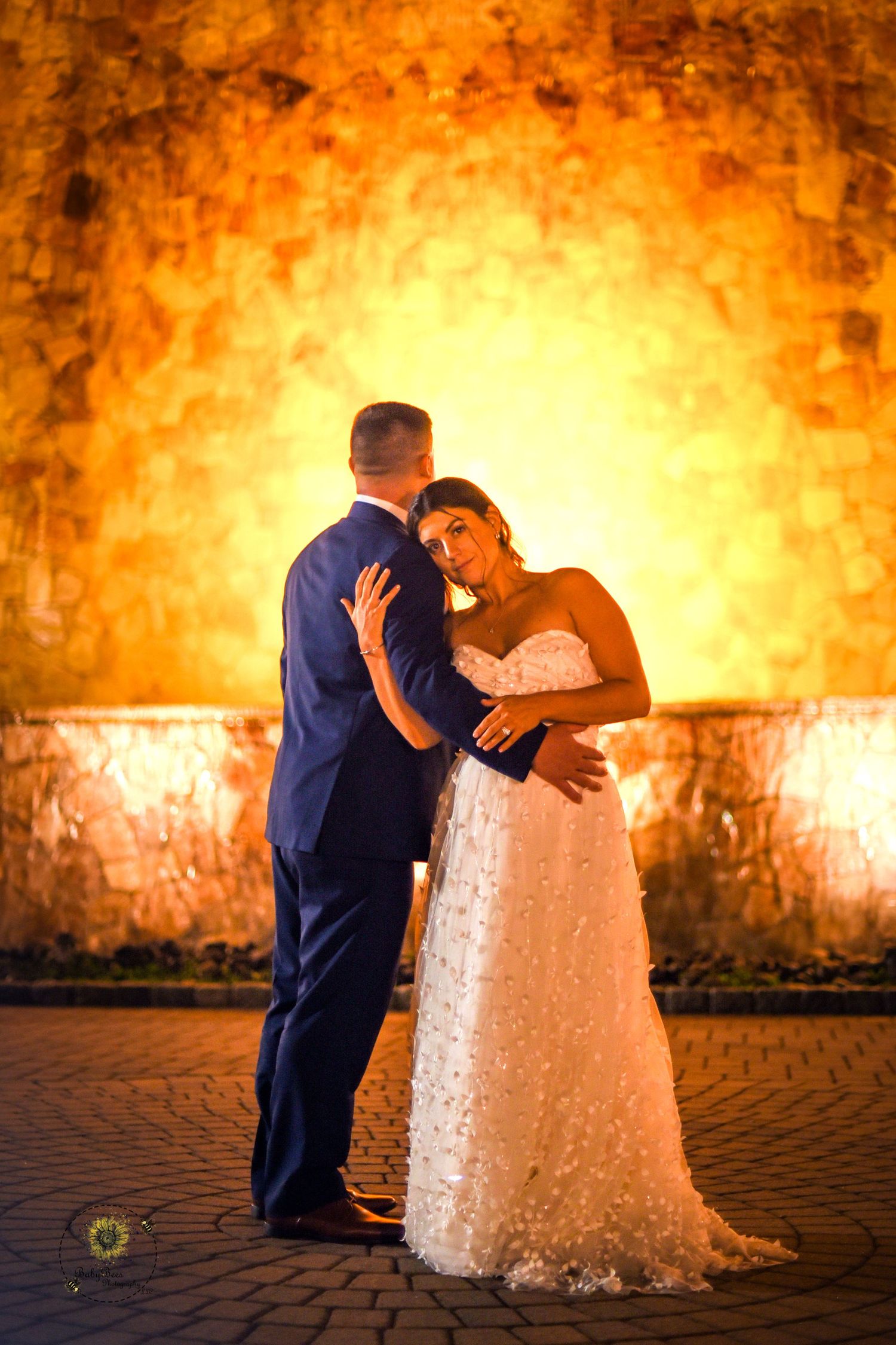 Romantic wedding couple embraces against a warm glowing stone wall backdrop at sunset.