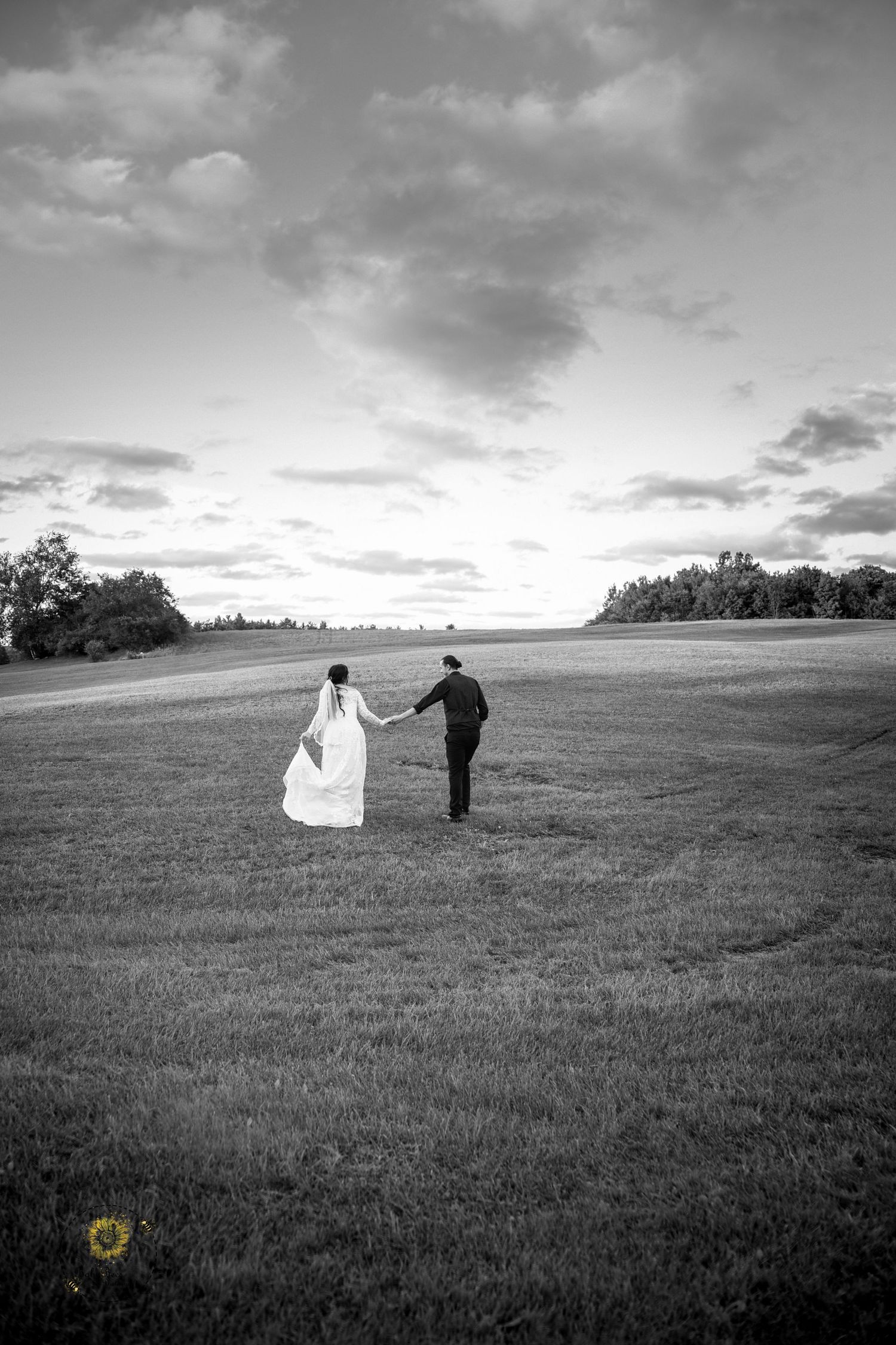 Black and white photo of newlyweds walking hand in hand across a vast open field.