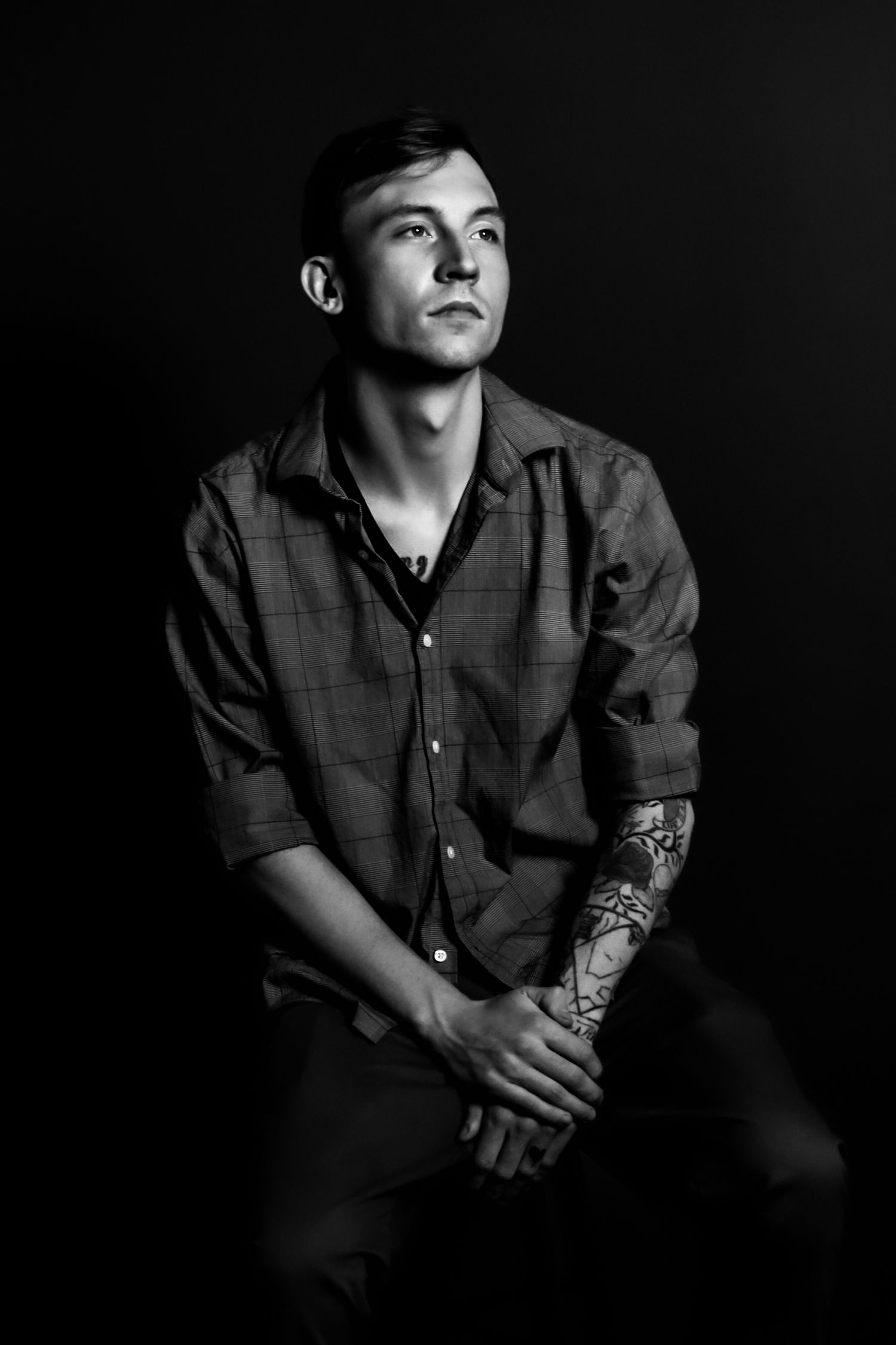 Black and white portrait of a person in a plaid shirt with a tattooed arm looking upward against a dark background.