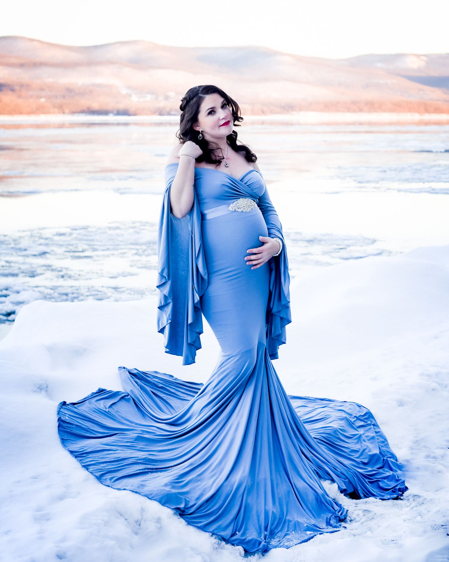 A flowing blue maternity dress billows dramatically against a snowy winter landscape at sunset.