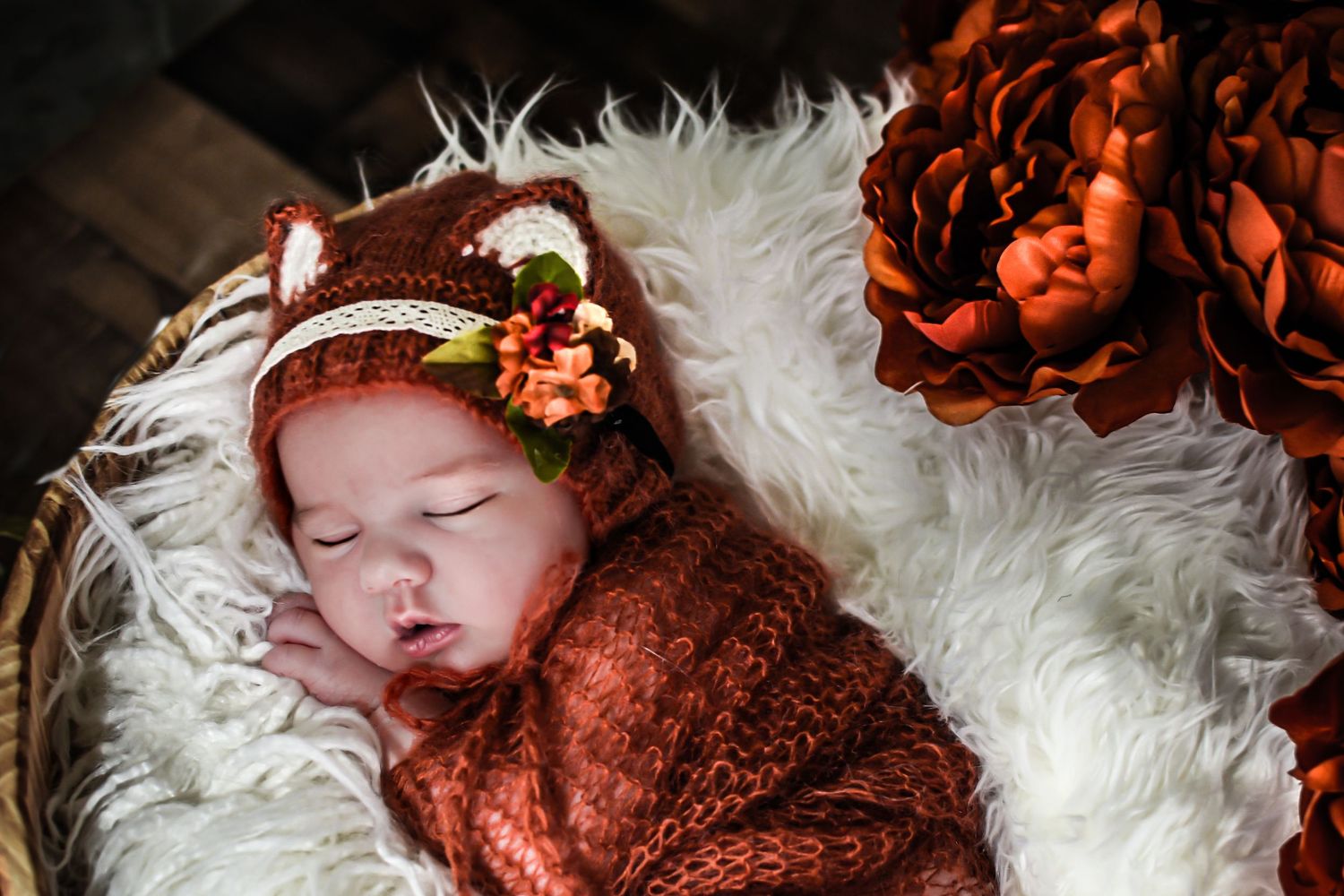 A sleeping newborn baby wearing a red outfit lies on soft white fur with red floral accessories.