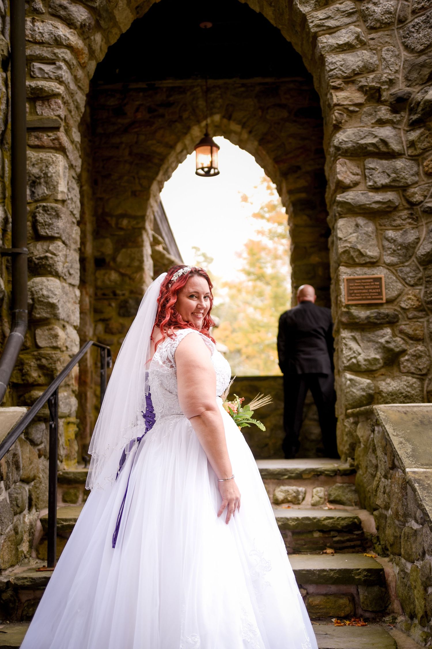 Bride in white gown poses under stone archway during fall season.