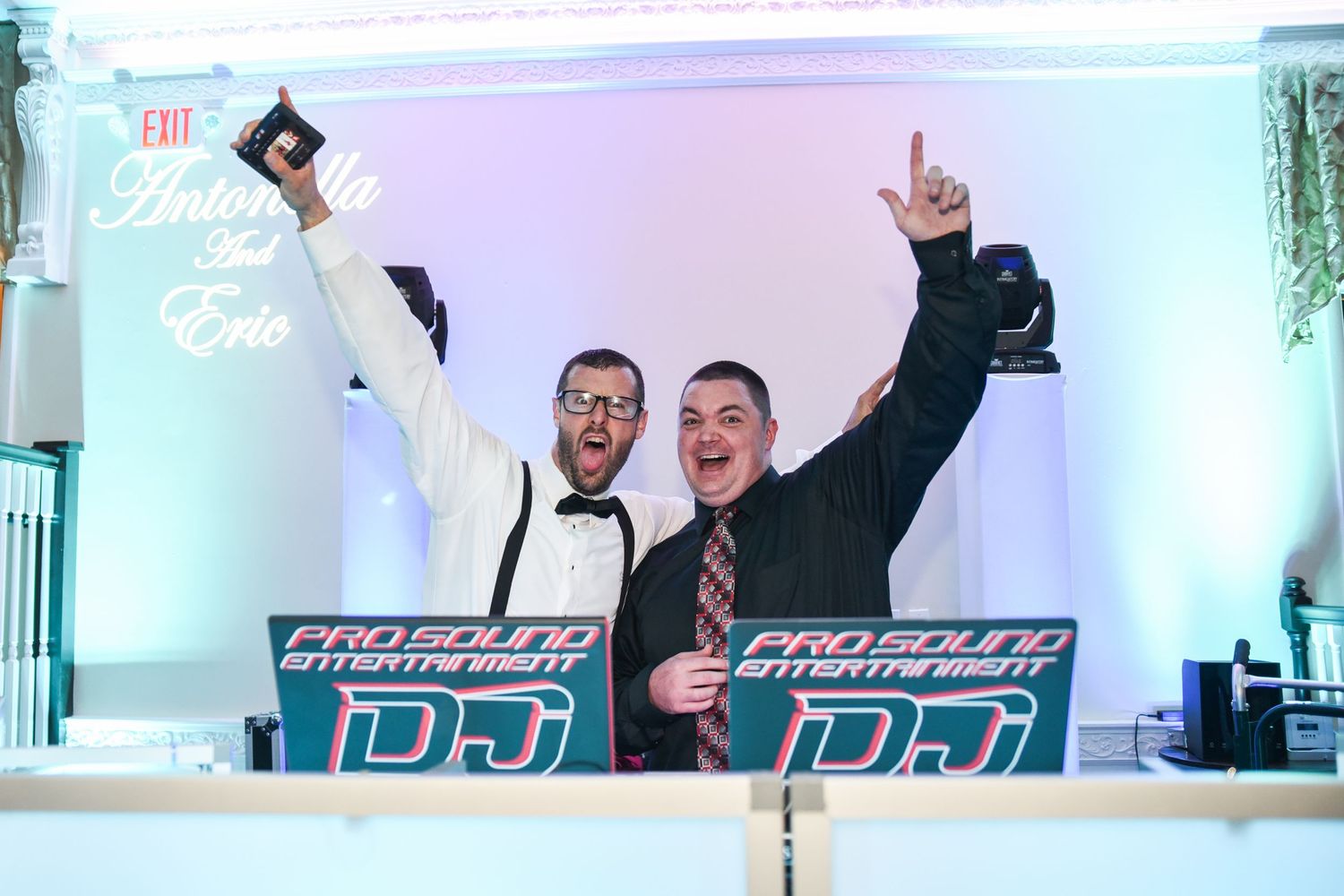 Two DJs performing energetically behind their booth at a wedding reception.