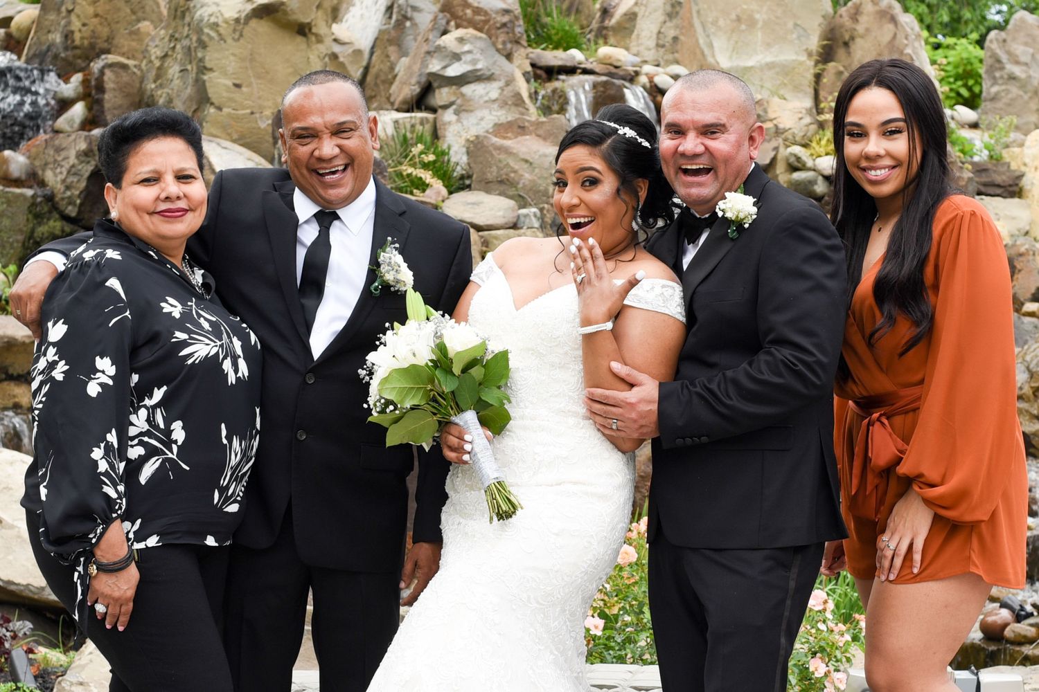 Wedding party poses together outdoors against stone wall backdrop with floral decorations.