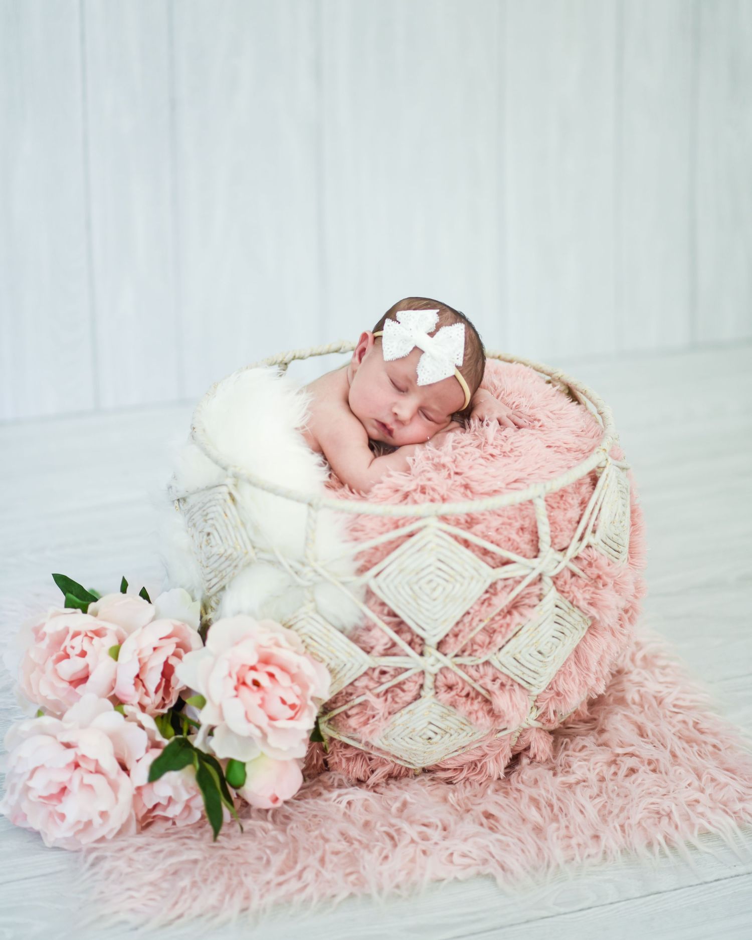A newborn sleeps in a white basket with pink fur and roses, creating a soft and dreamy newborn photography setup.