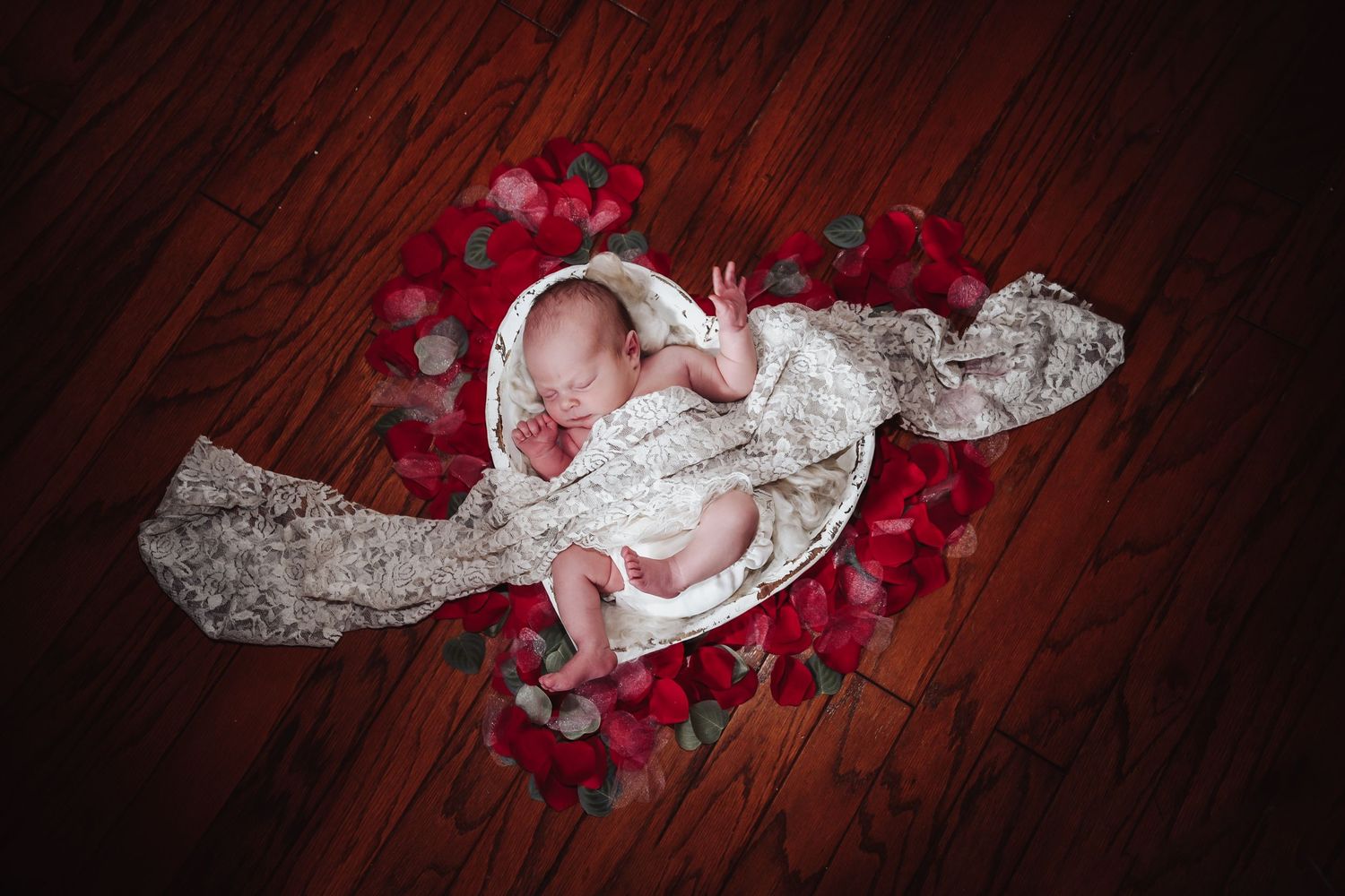 White lace wedding dress and red rose petals scattered on a dark wooden floor in romantic lighting.