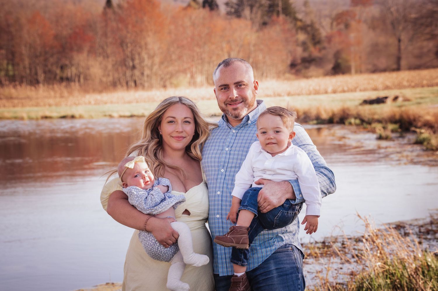 A family stands together outdoors near a lake during autumn with golden grass and trees in the background.