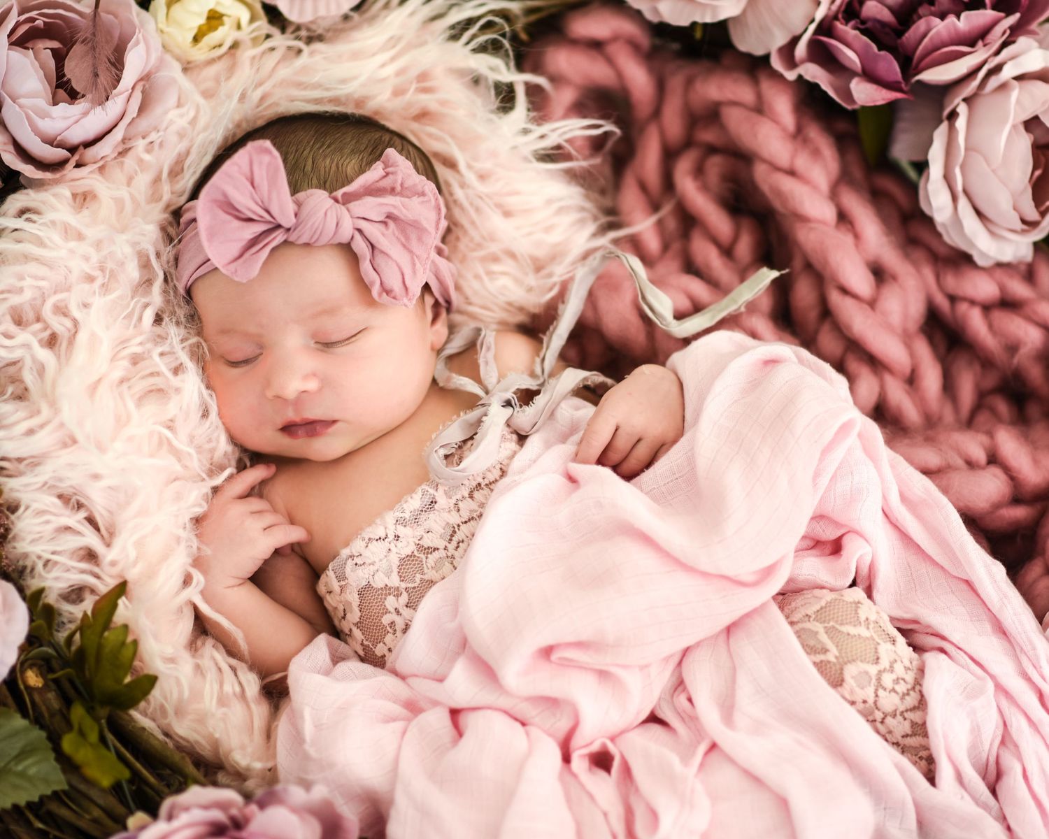 A sleeping baby wearing a pink floral headband rests peacefully among soft pink and white roses.