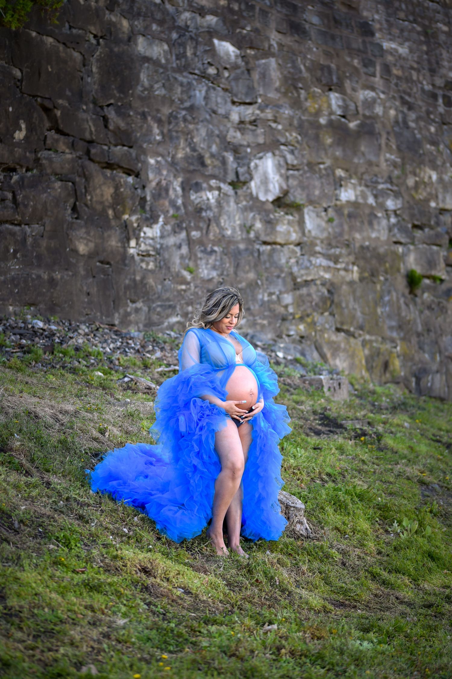 A figure in a vibrant blue tulle dress sits thoughtfully against an ancient stone wall on a grassy slope.