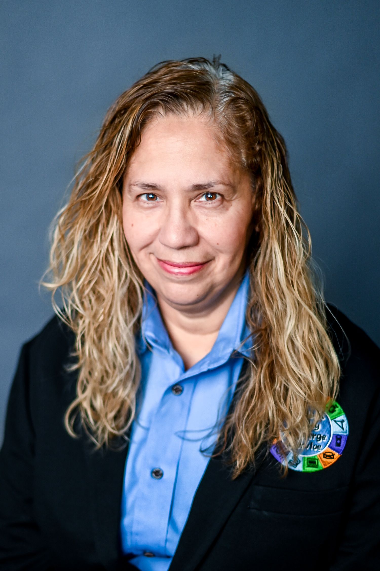 Professional headshot portrait with wavy blonde hair wearing a black blazer and blue button-up shirt against dark background.