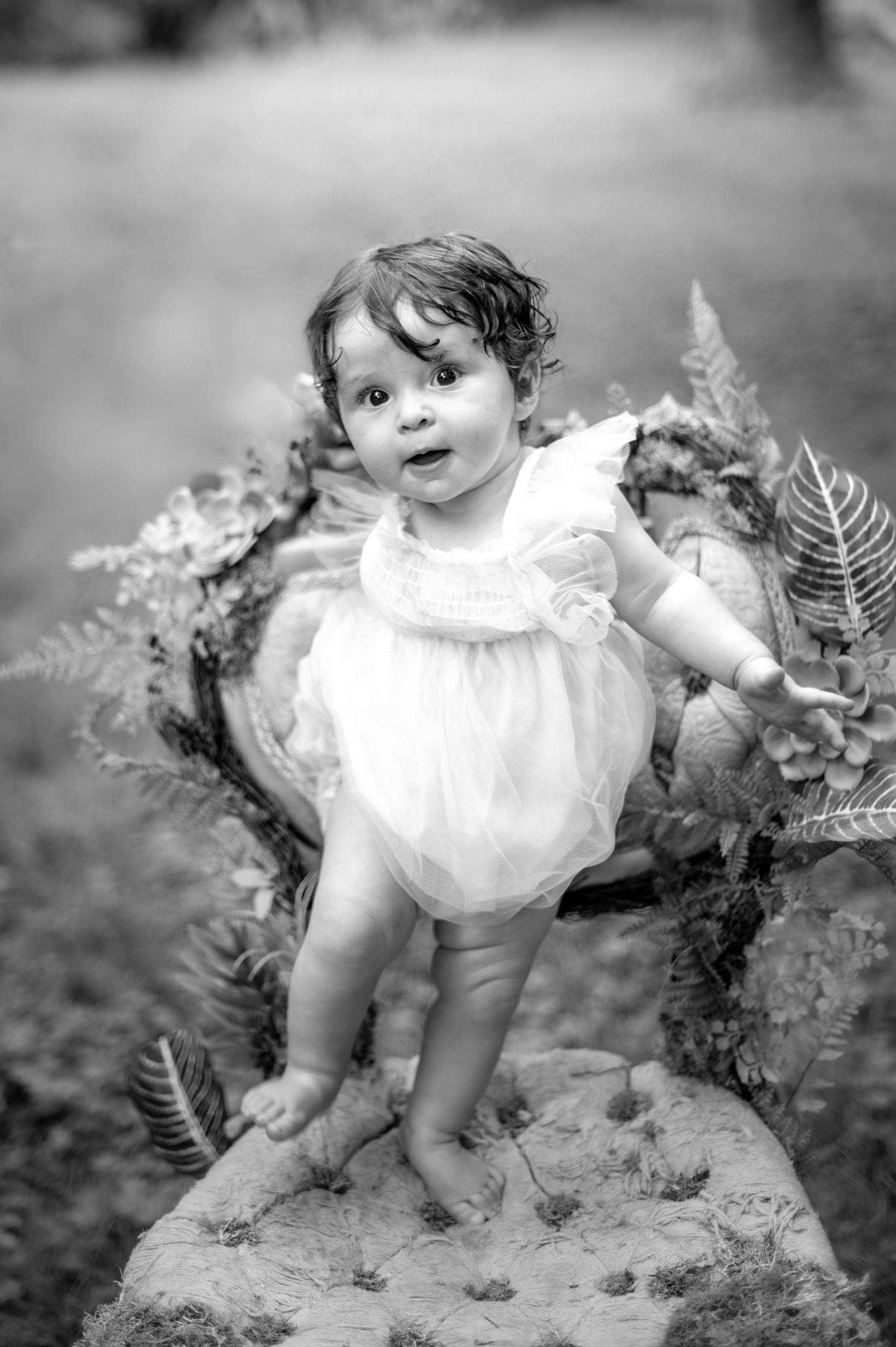 Sweet toddler in white dress standing on log surrounded by ferns in black and white.