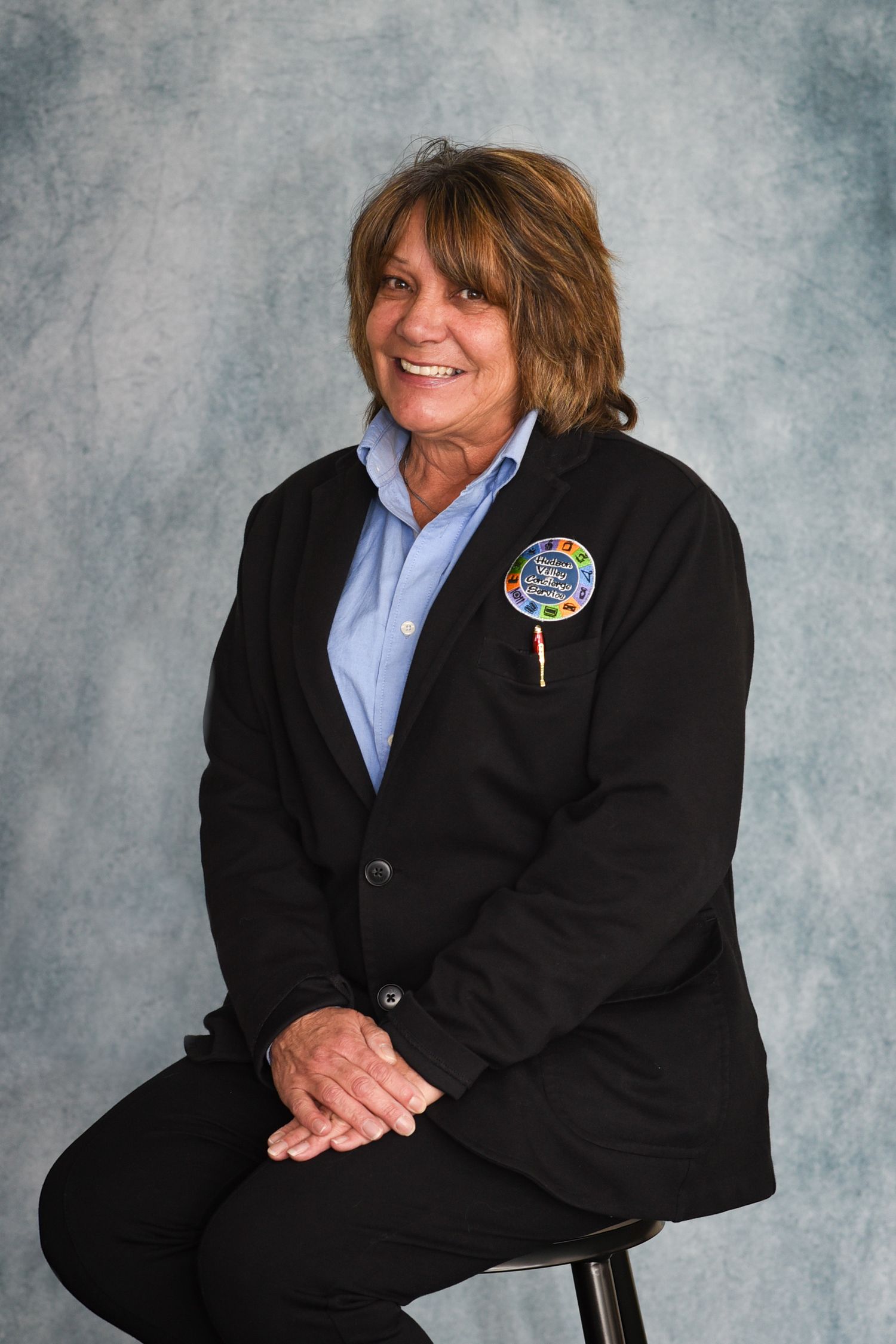 Professional studio portrait of someone in a black blazer and blue shirt with a decorative pin, seated on a stool.