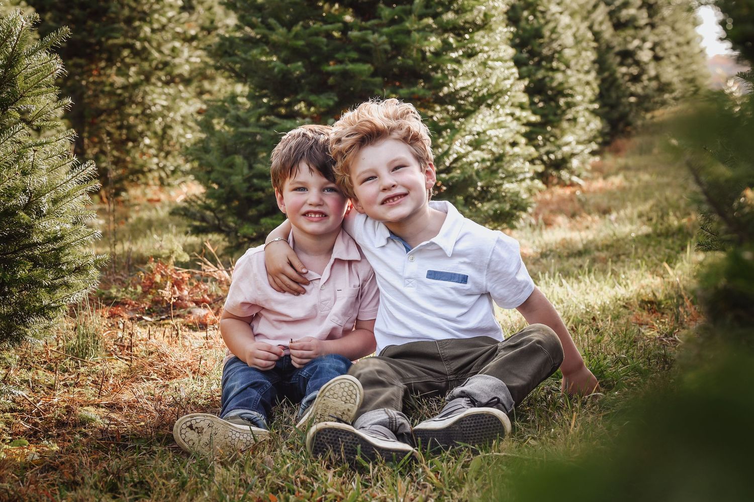 Two children sit together on autumn ground surrounded by evergreen trees in a natural outdoor setting.
