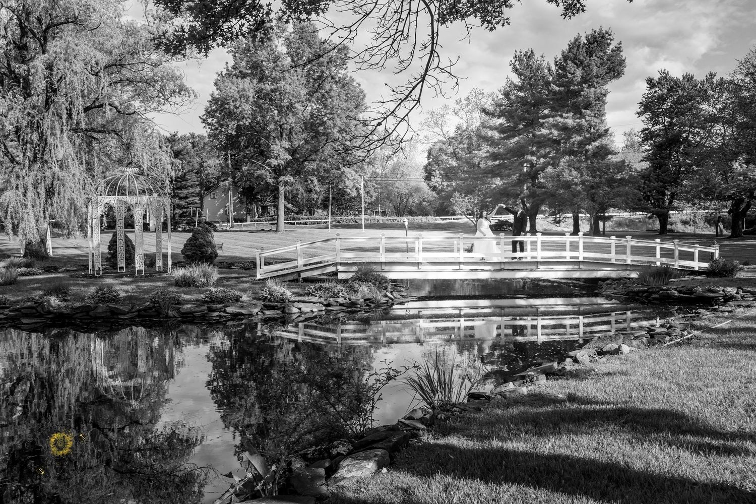 A scenic black and white photograph of a bridge over a pond with reflections in the water.