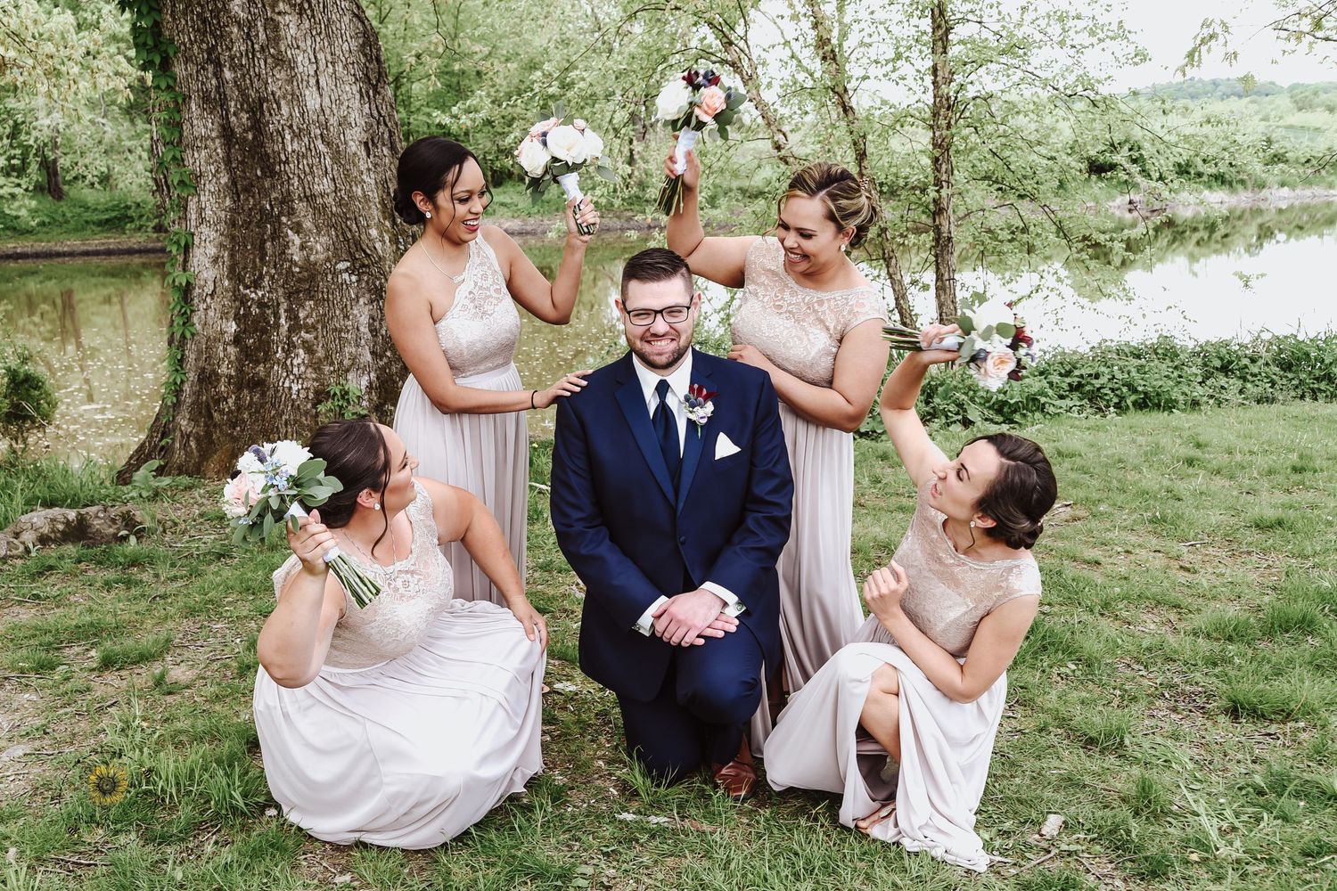 Wedding party poses playfully on grass near a large tree.