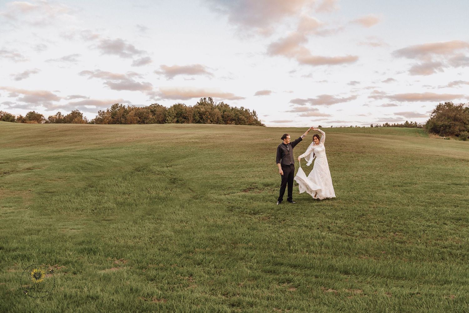 A wedding couple stands in a scenic field during golden hour with dramatic clouds in the background.