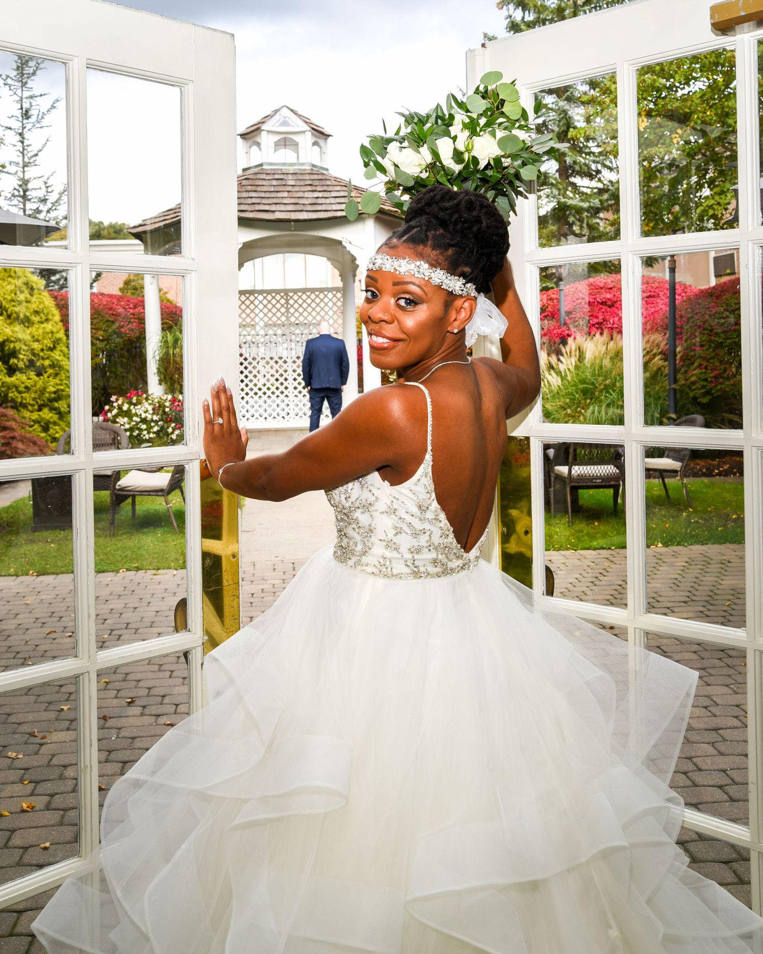 Bride in elegant white ball gown poses playfully by French doors in garden setting.