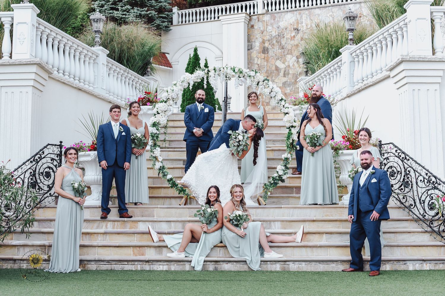 Wedding party poses on grand white stone staircase during outdoor ceremony.