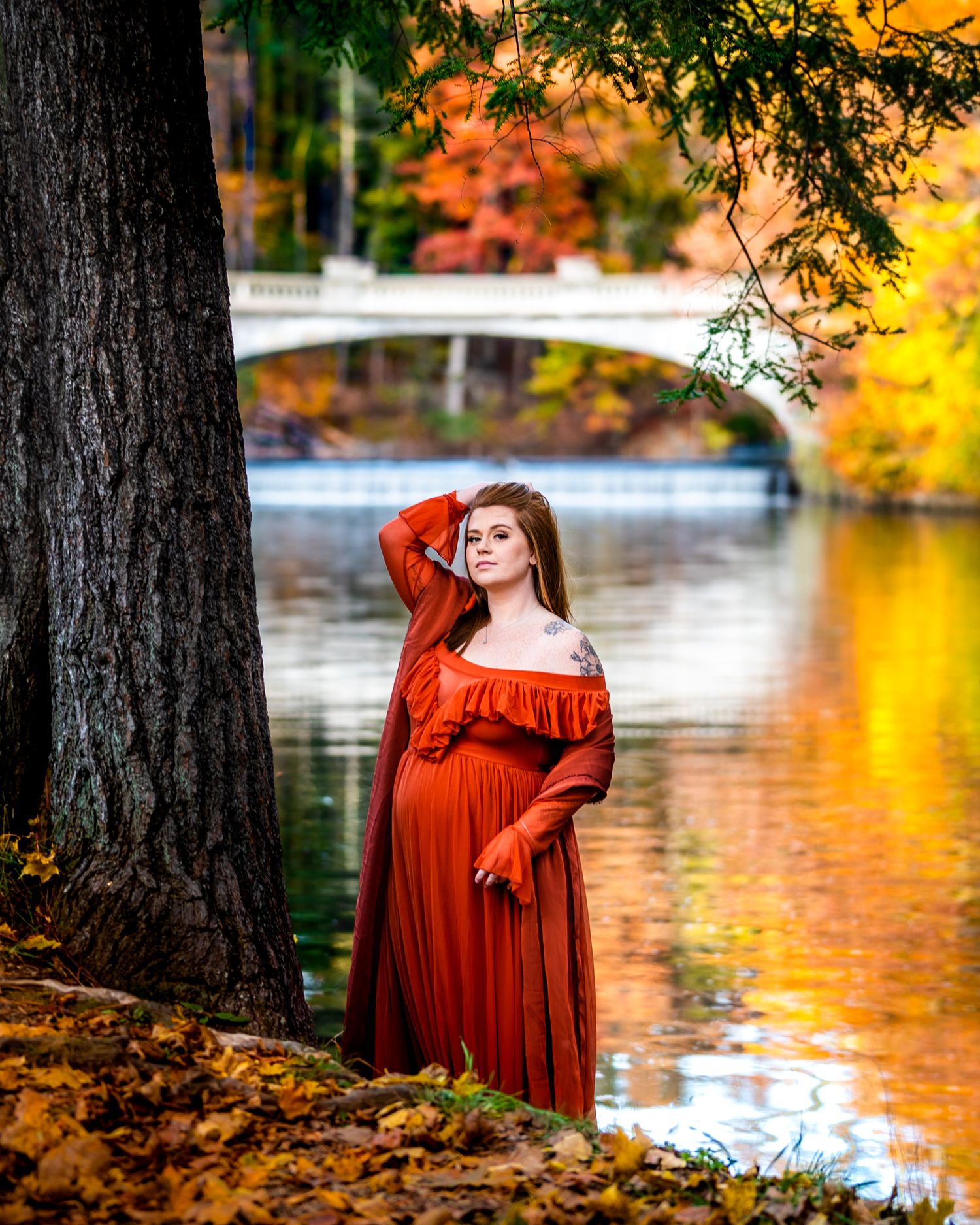A person in a flowing red dress poses gracefully by a lake surrounded by autumn foliage and golden light.